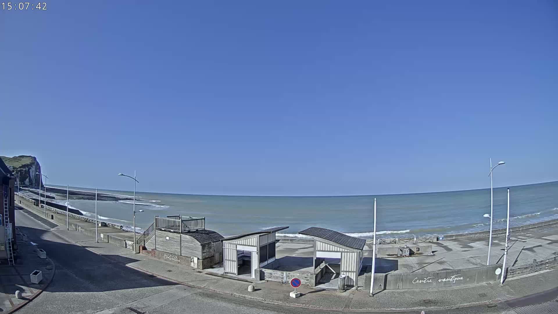 A mostly sunny day at a beach with calm waves, featuring a paved beachfront area, two small structures, and a distant cliff.
