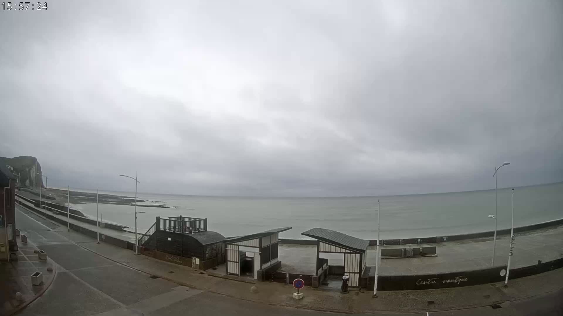 A mostly cloudy day shows an overcast sky above a calm sea with a rocky shoreline and a paved road leading to the water's edge, next to which are some structures that appear to be beach access points.