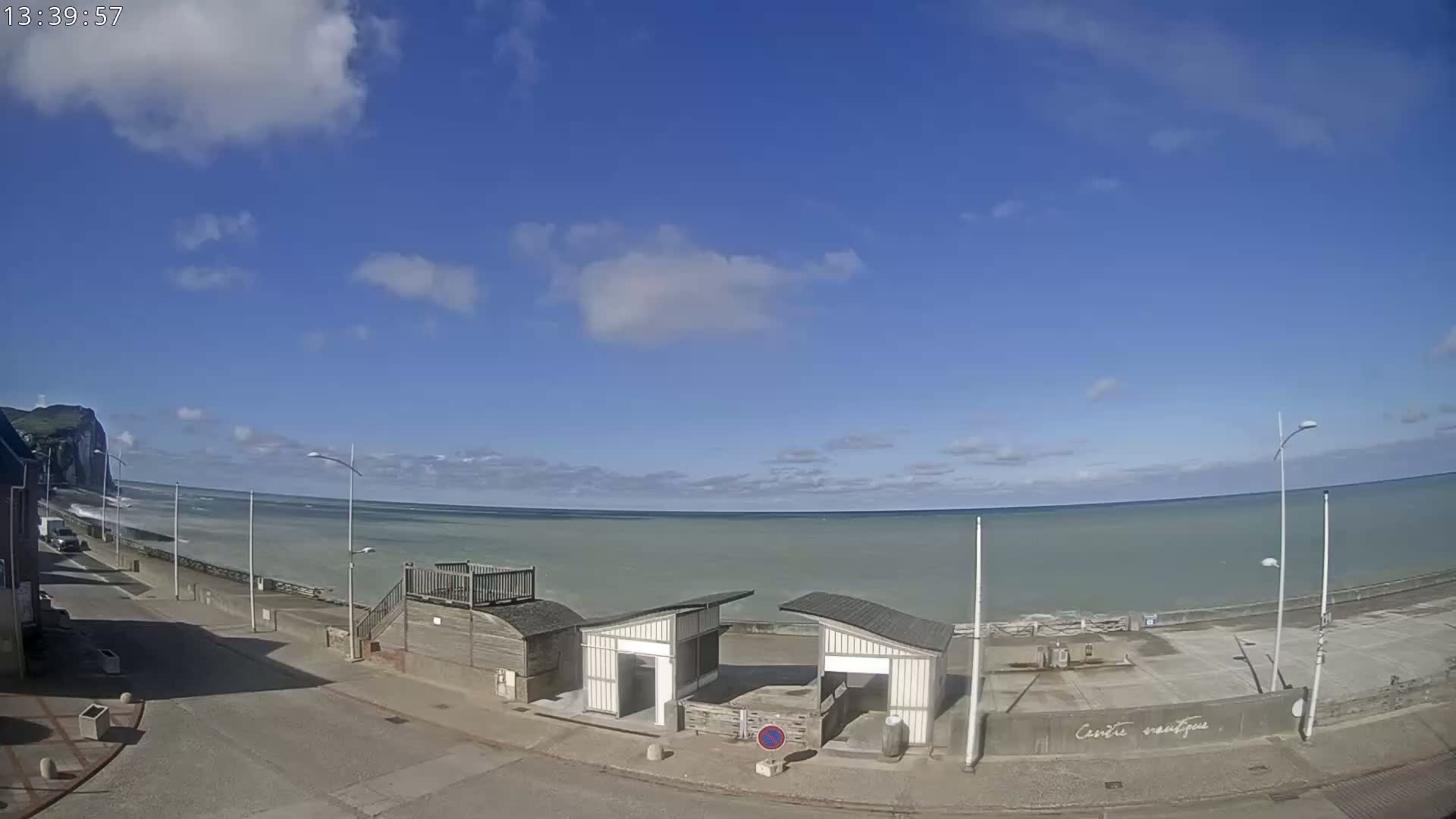 A partly cloudy blue sky overlooks a calm, light-green ocean meeting a paved coastal area with small structures and a distant white cliff.