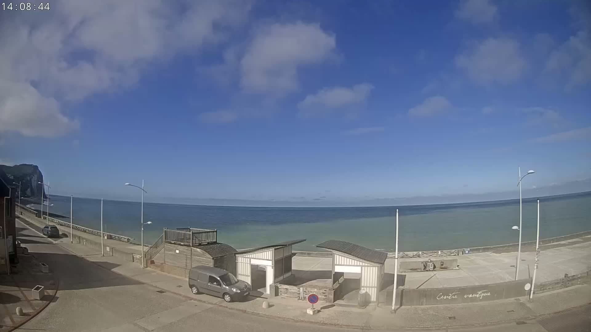 A mostly sunny day overlooks a calm ocean, with a beach, buildings, and a van visible from a coastal street.