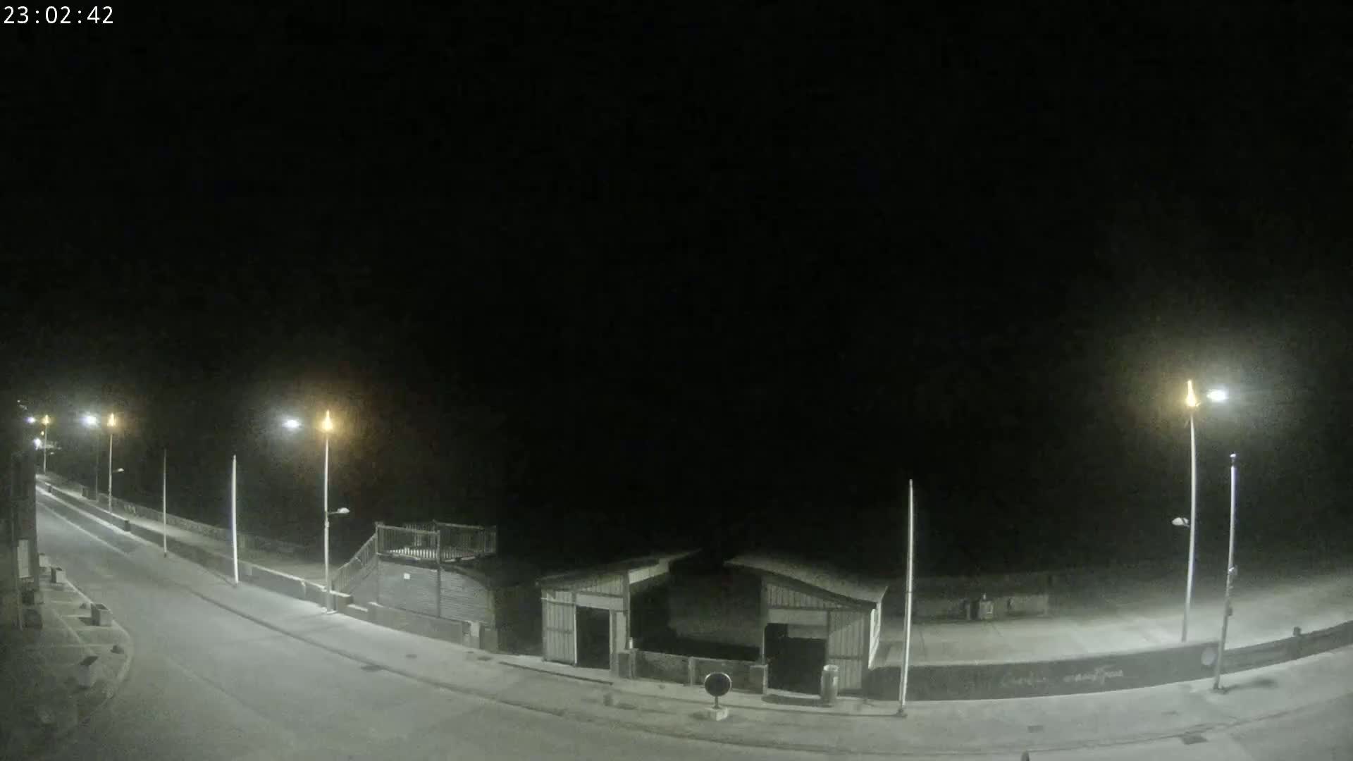 A mostly empty, paved road curves along a beachfront at night, illuminated by streetlights under a dark sky.