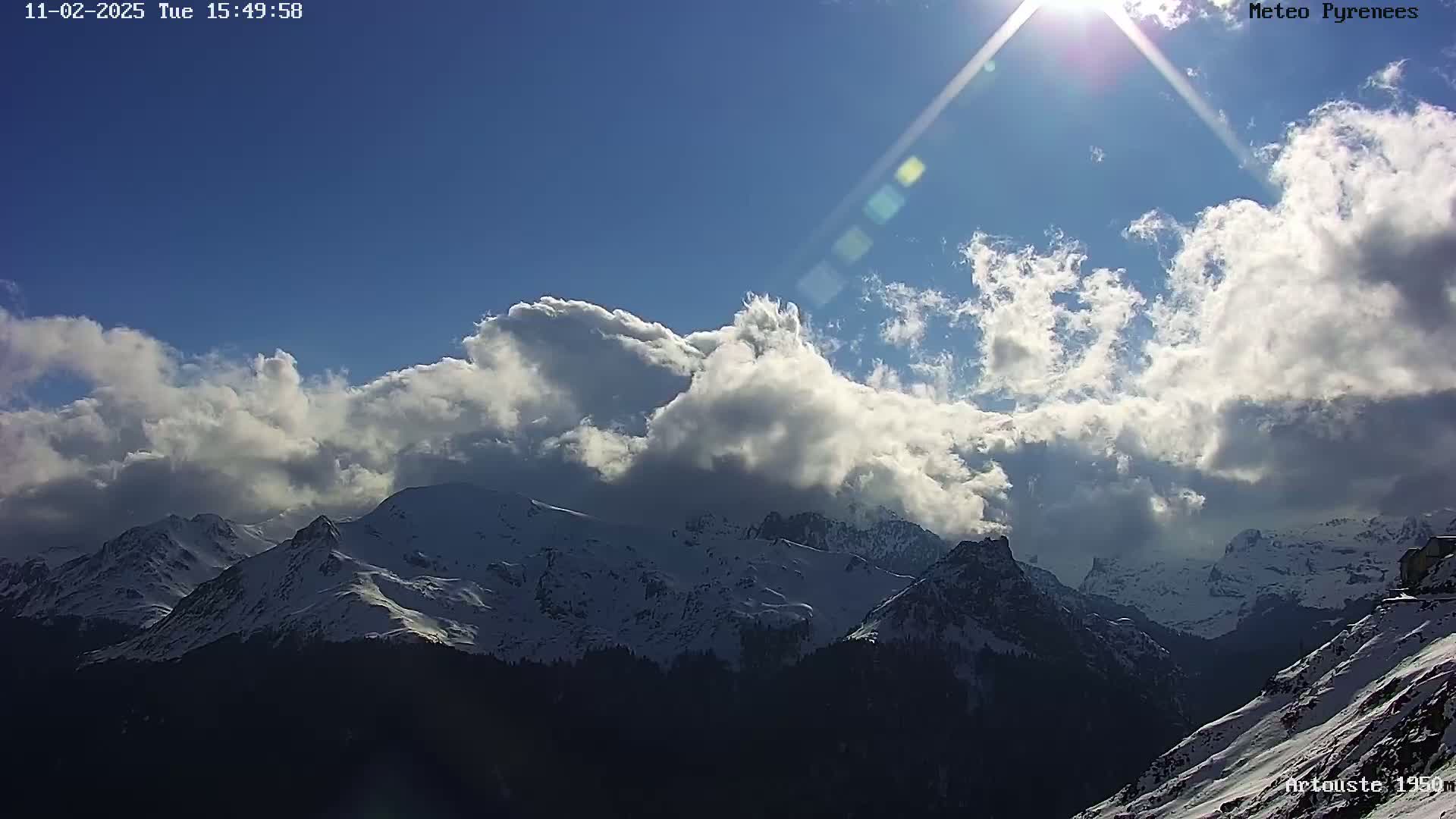 A mostly sunny day shows a range of snow-capped mountains partially obscured by fluffy white clouds.
