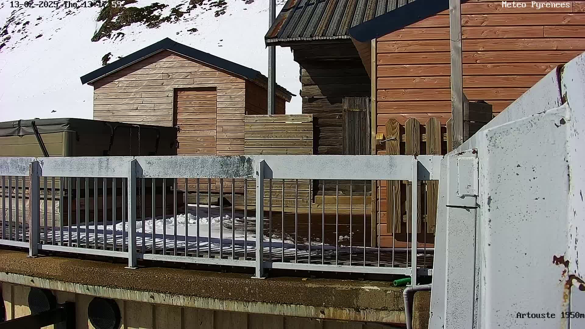 A metal railing overlooks a snowy landscape with several wooden structures, including small cabins and what appears to be a hot tub.