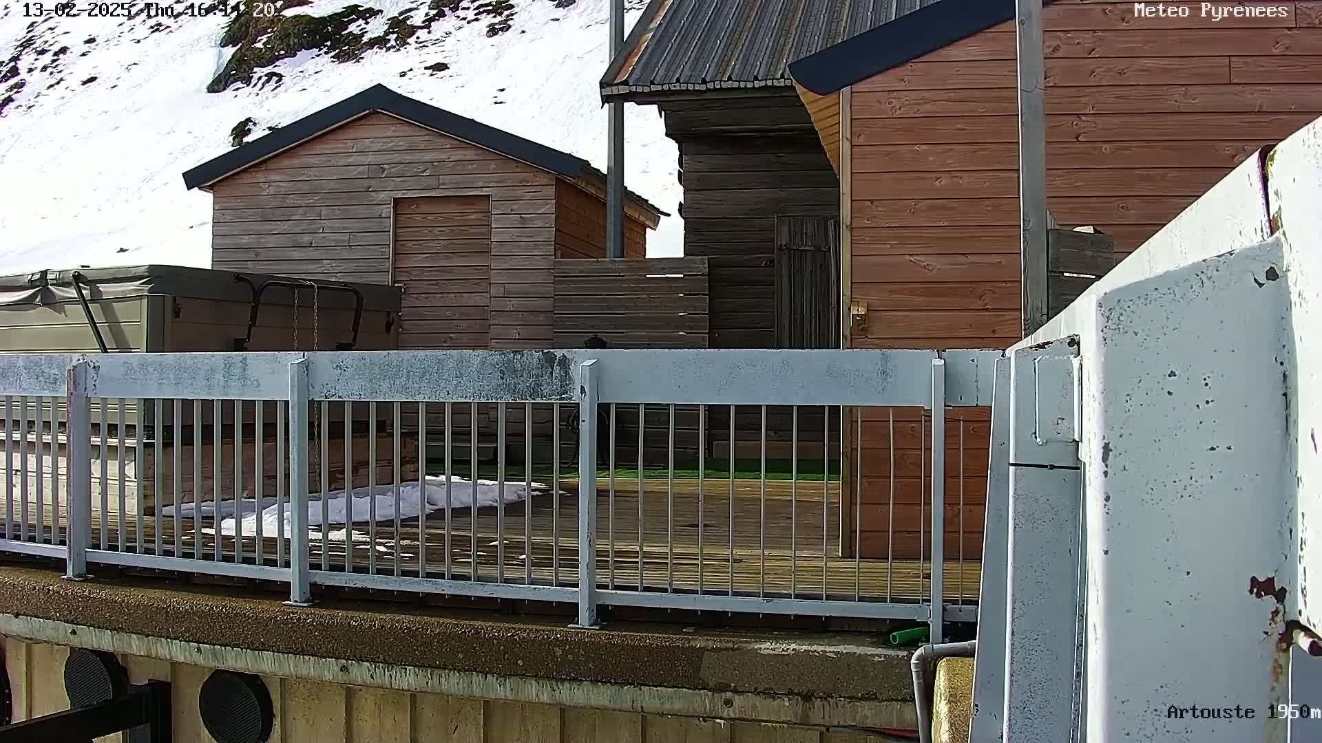 A deck with a hot tub and several wooden buildings is partially visible behind a metal railing, with some snow on the ground and a snowy hillside in the background.