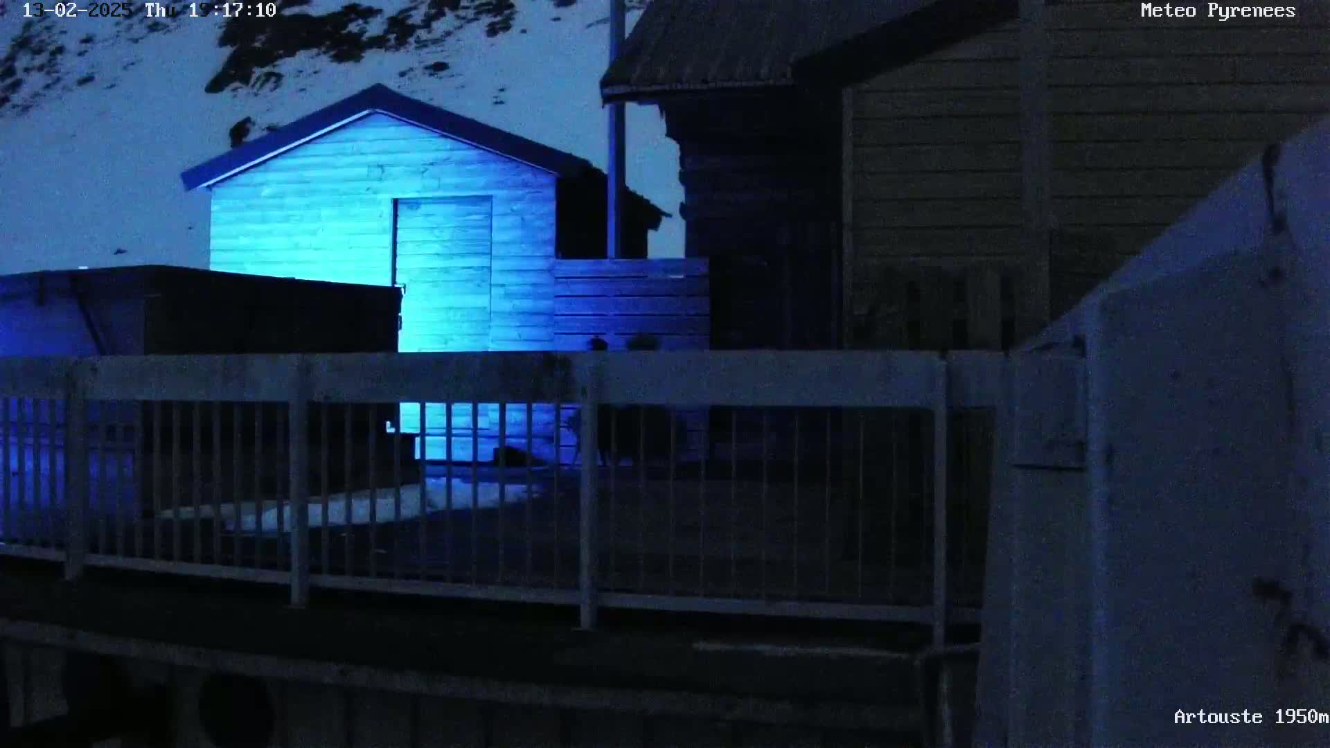 A nighttime scene shows a small, blue-lit shed and other dark buildings behind a metal fence in snowy conditions.