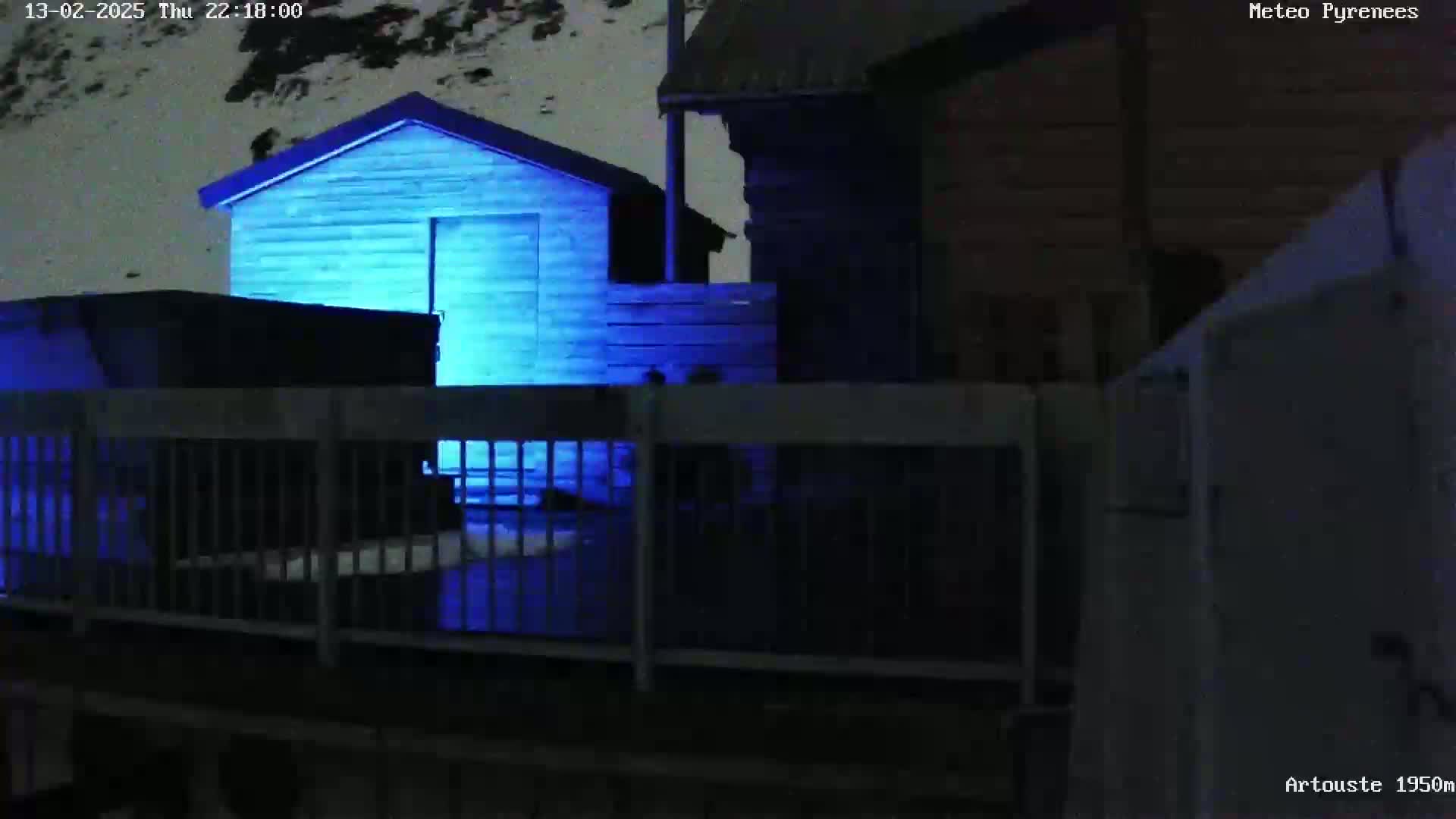 A small, blue-lit shed sits behind a fence in a snowy, nighttime scene near other dark buildings.
