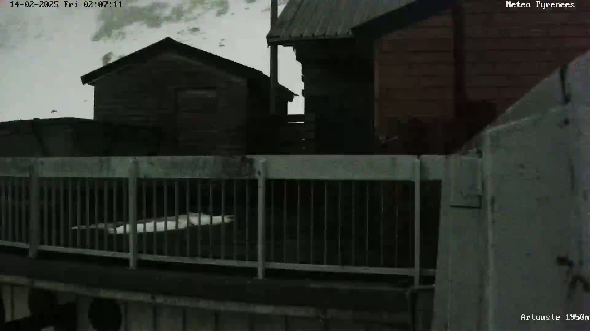 Snow falls on dark wooden buildings and a metal fence viewed from a slightly elevated perspective.