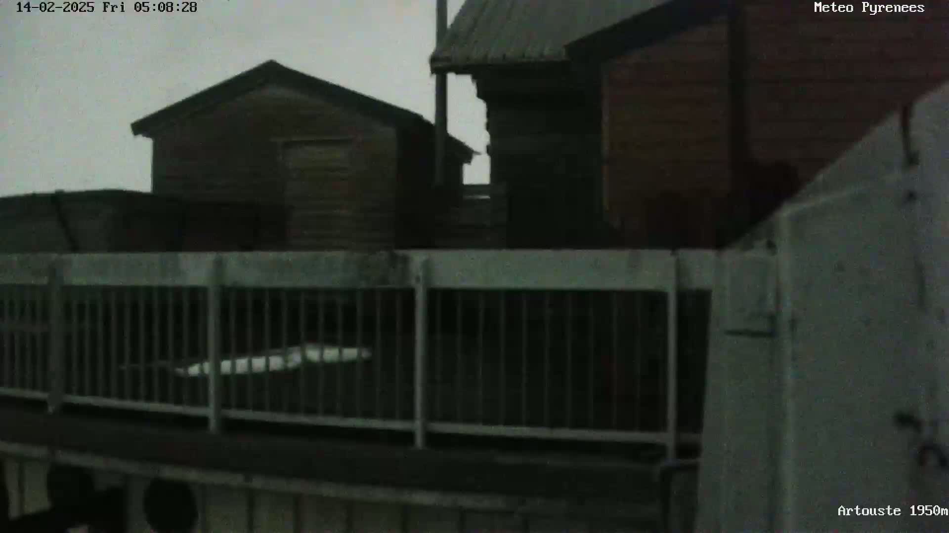 A view from behind a metal railing shows several dark-colored wooden buildings under an overcast sky.