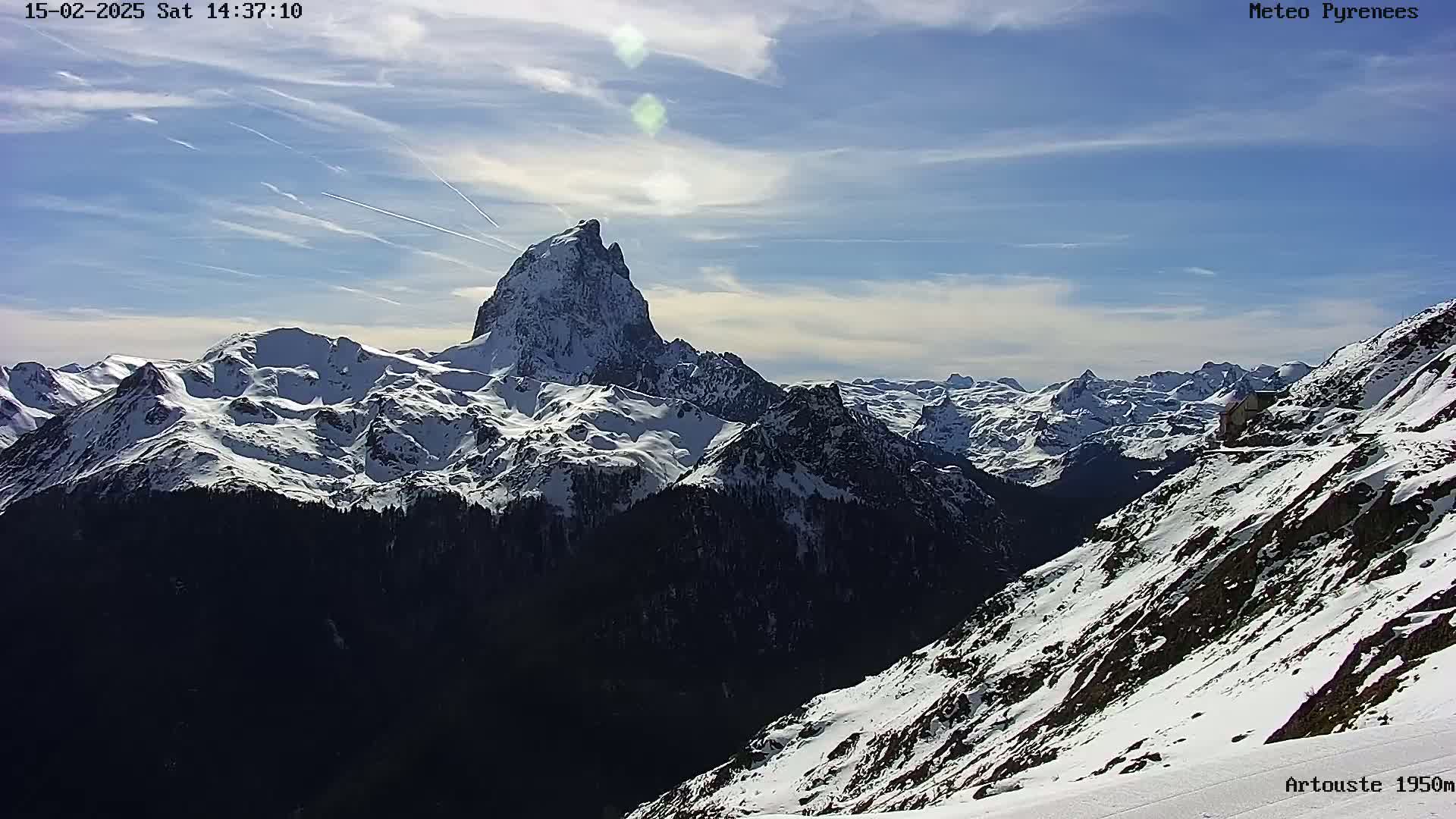 A snow-capped mountain range under a mostly clear, sunny sky with contrails visible.