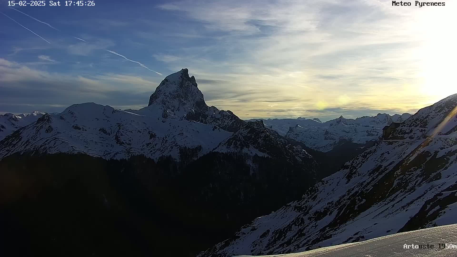 A snow-covered mountain range is illuminated by the setting sun under a partly cloudy sky.