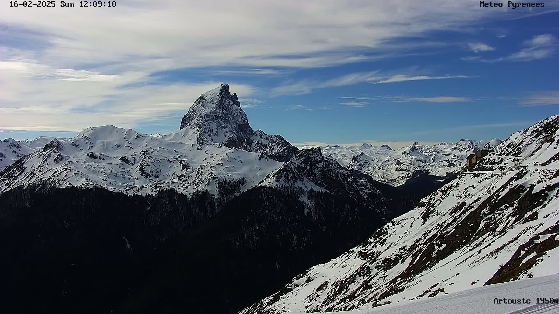 A snow-covered mountain range with a prominent, sharply peaked mountain in the center, under a mostly clear, bright blue sky with some wispy clouds.
