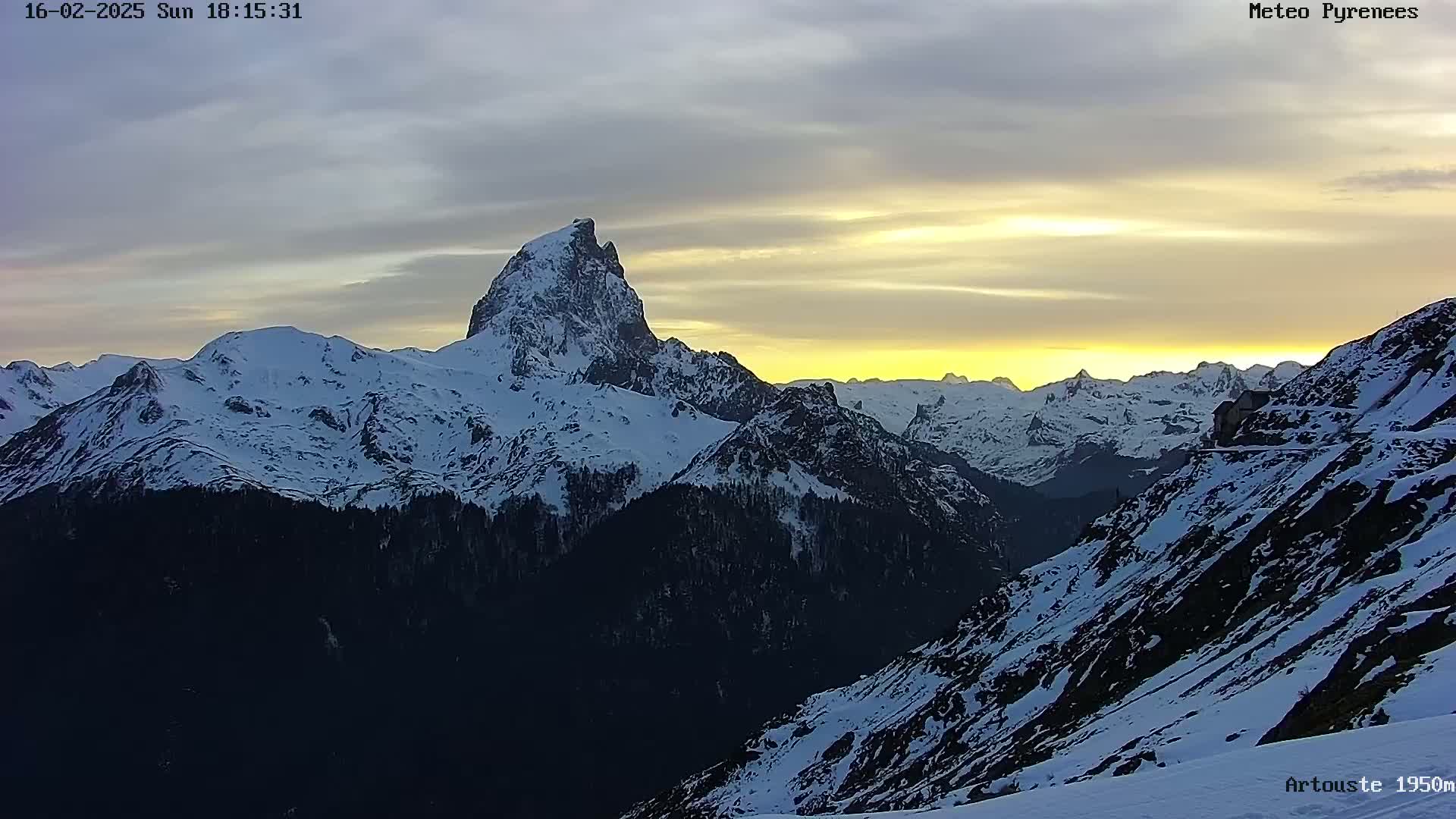 A snow-covered mountain range is illuminated by a setting sun under a mostly clear sky.