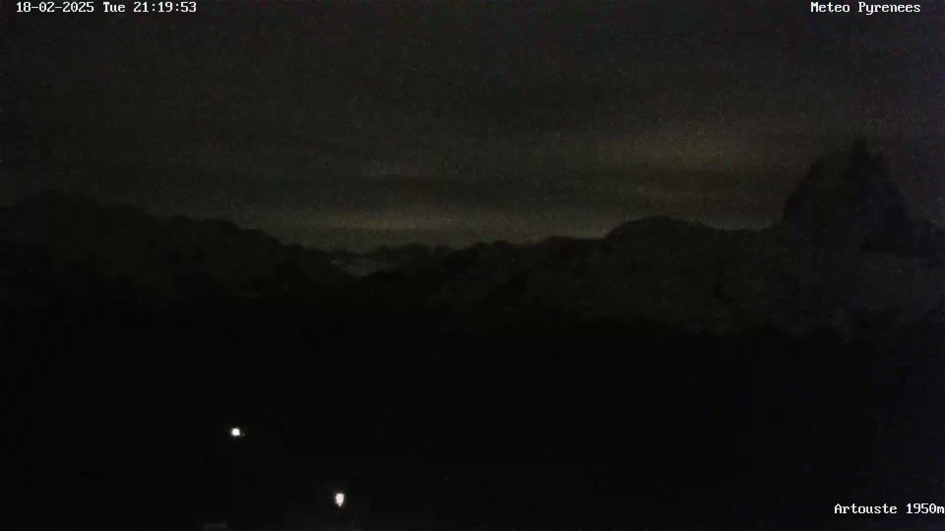 A nighttime, dark view of silhouetted mountains under a mostly clear sky with a few faint clouds.
