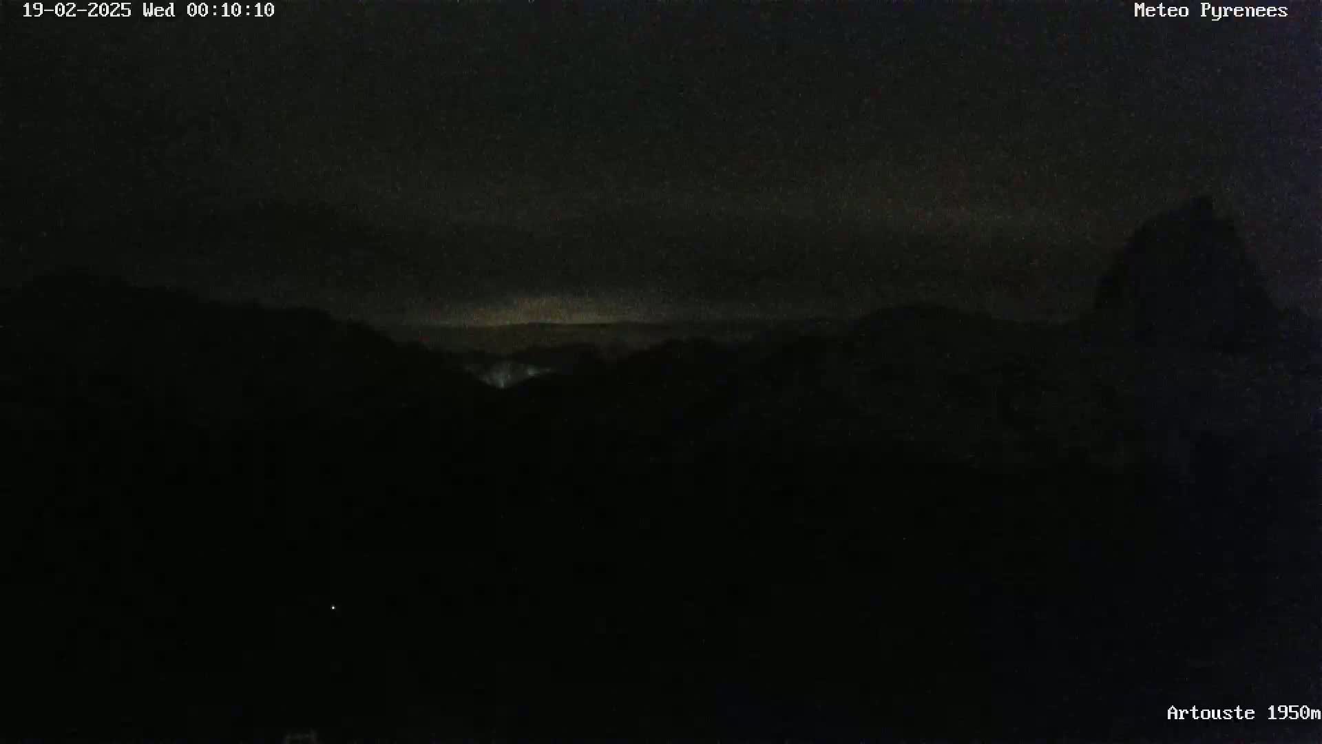 A nighttime, dark landscape of mountains is shown under a cloudy sky with a faint light on the horizon.