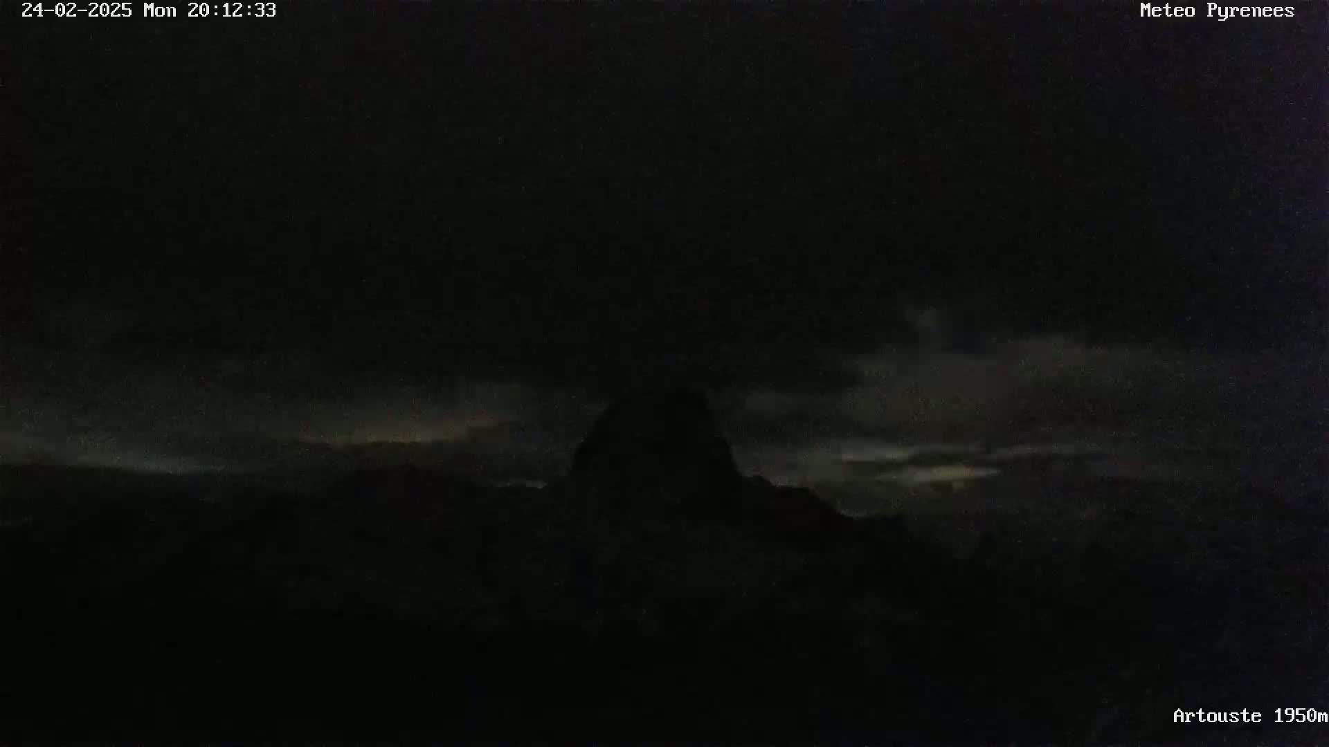 A dark, nighttime landscape shows a silhouetted mountain peak against a faintly illuminated sky with clouds.