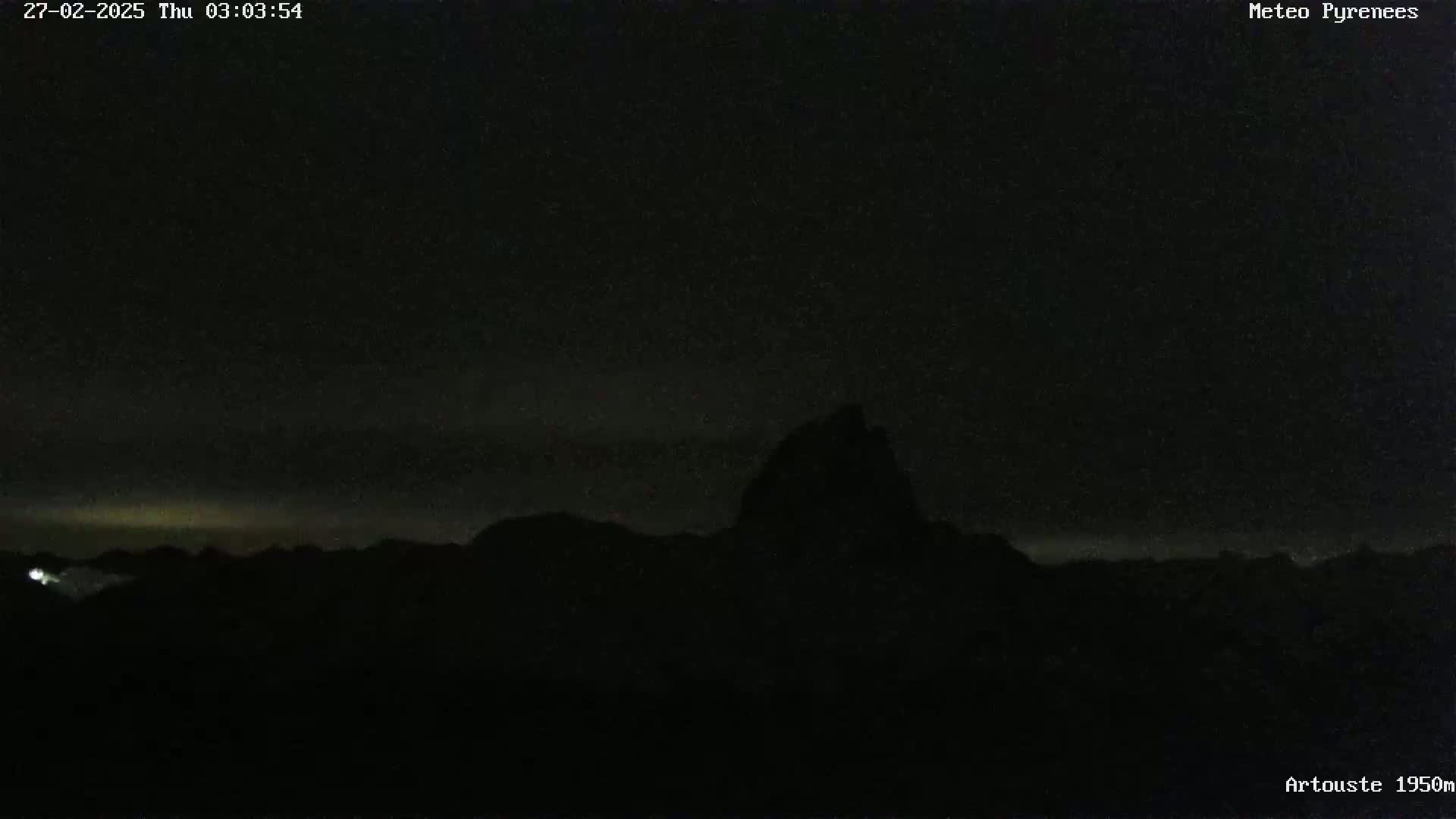 A dark, nighttime landscape shows a silhouetted mountain range under a mostly clear sky with a faint, light horizon.