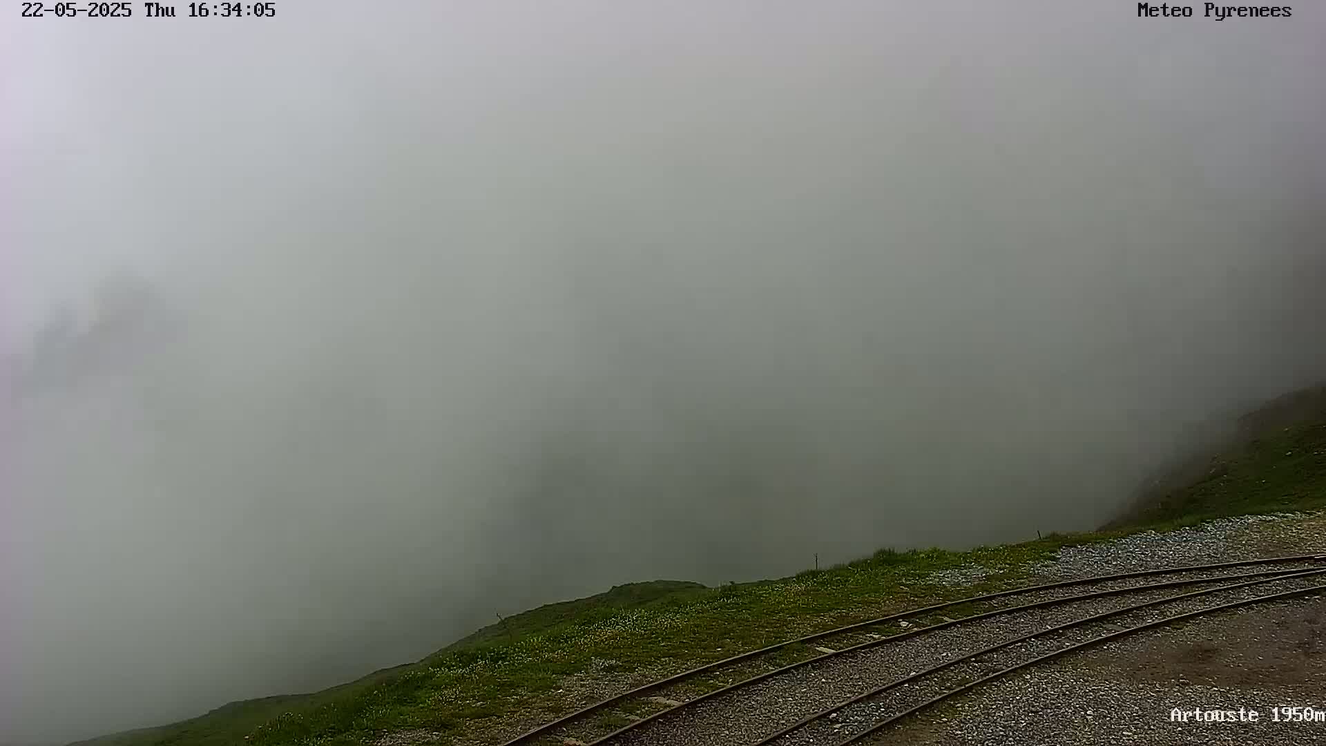 A foggy, mountainous landscape is seen from a high vantage point, showing train tracks curving alongside a grassy hillside.