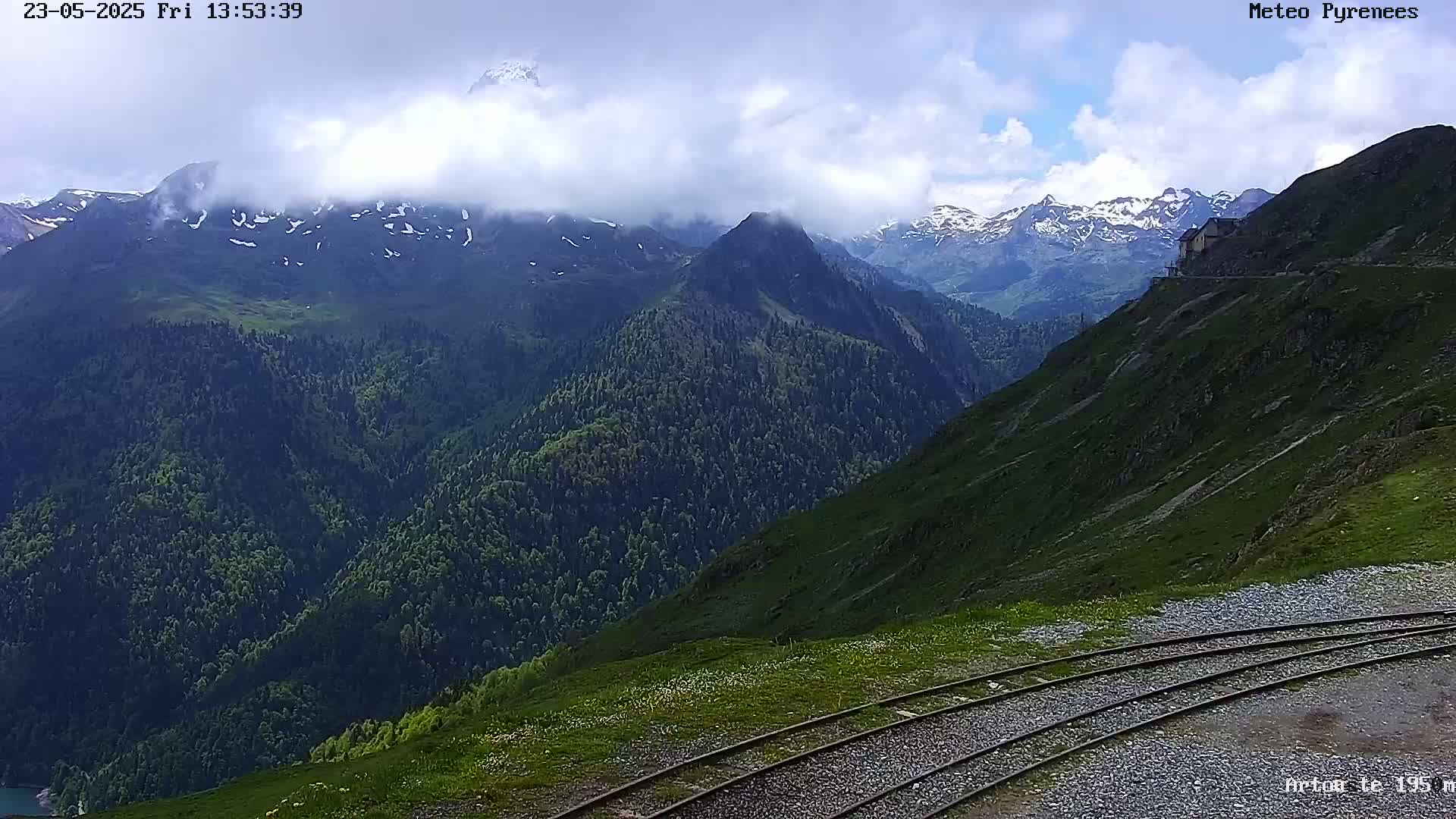 A partly cloudy day reveals a mountain range with snow-capped peaks, partially obscured by clouds, and a train track curving along a grassy hillside.