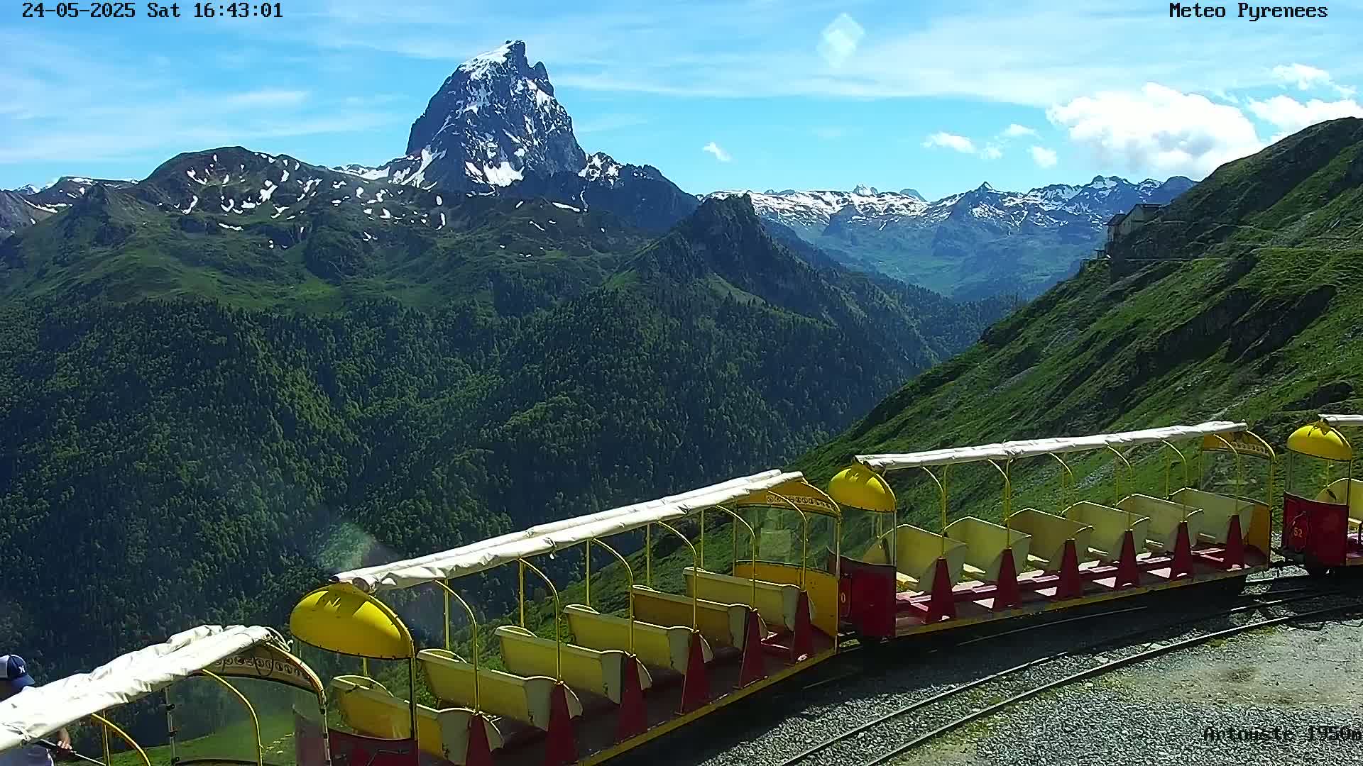 A yellow and red train sits on a mountainside overlooking a valley and a snow-capped mountain peak under a mostly sunny sky.