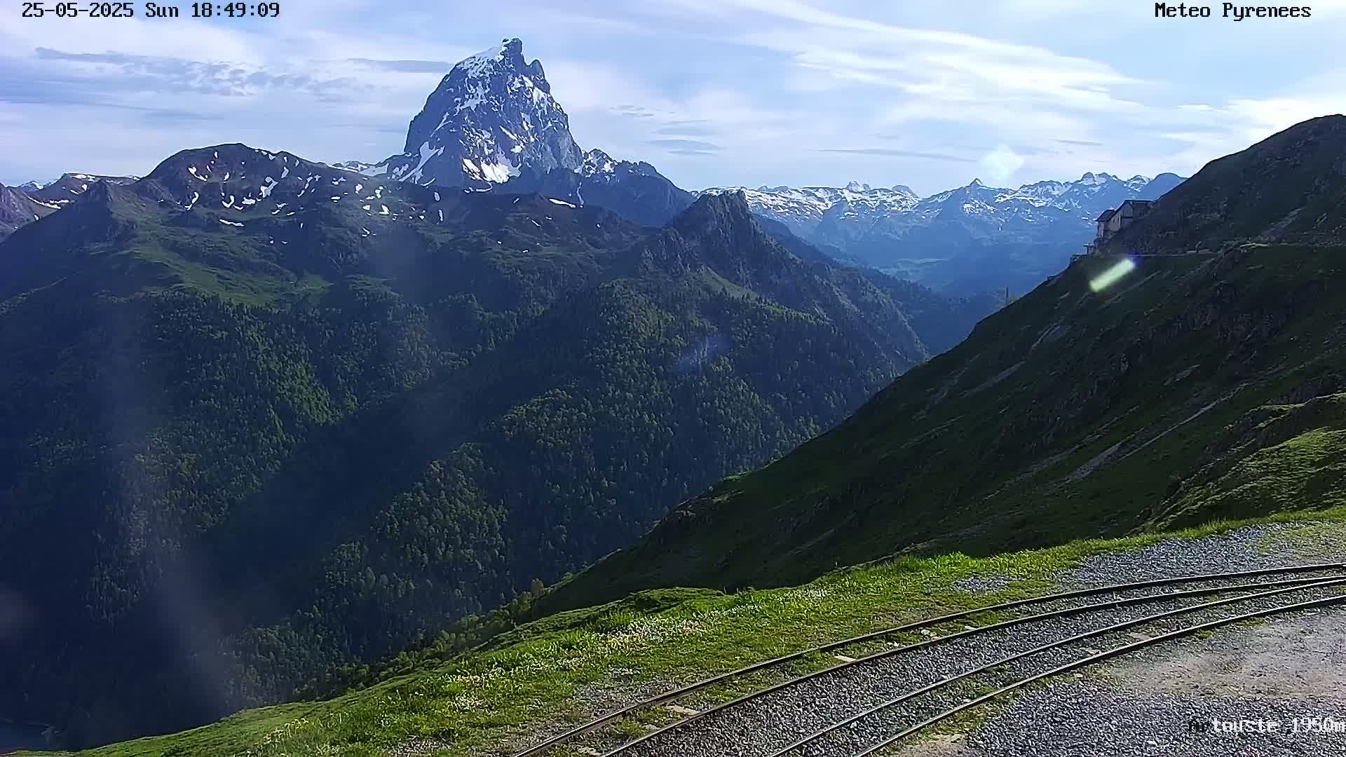 A mountain range with a prominent snow-capped peak is visible under a mostly clear sky, with train tracks in the foreground.