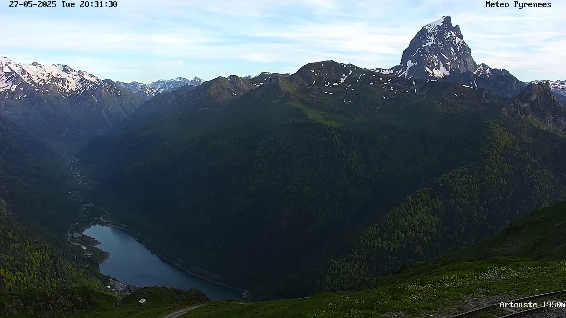 A mountain lake nestled in a valley surrounded by dark green mountains, some with snow-capped peaks, under a mostly clear sky.