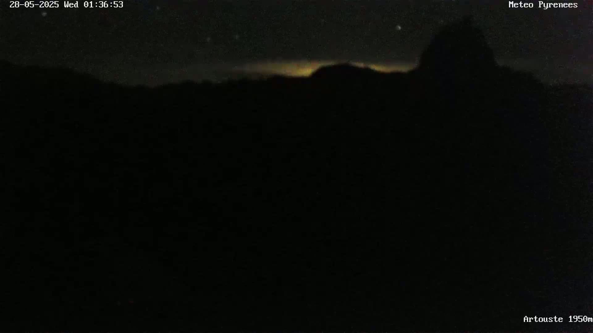 A dark, nighttime landscape shows a silhouetted mountain range under a mostly clear, starry sky with a faint, light-colored glow on the horizon.