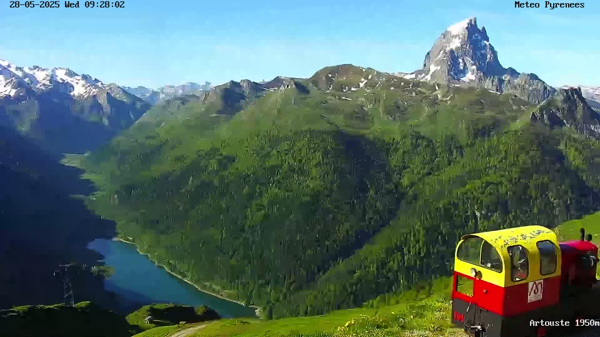 A small, red and yellow train is parked on a hillside overlooking a lake and a mountain range under a clear blue sky.