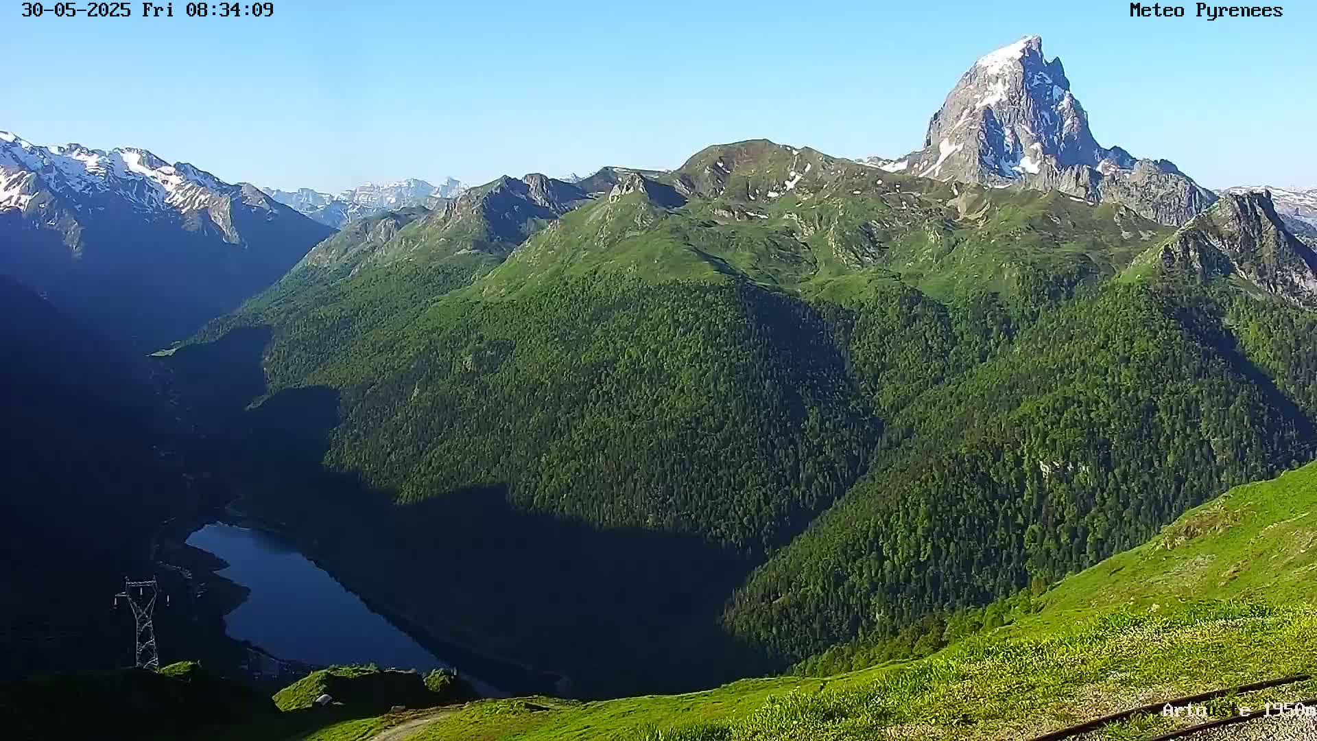 A clear, sunny day reveals a valley of lush green mountains and a dark blue lake, with a prominent snow-capped peak rising above the rest.