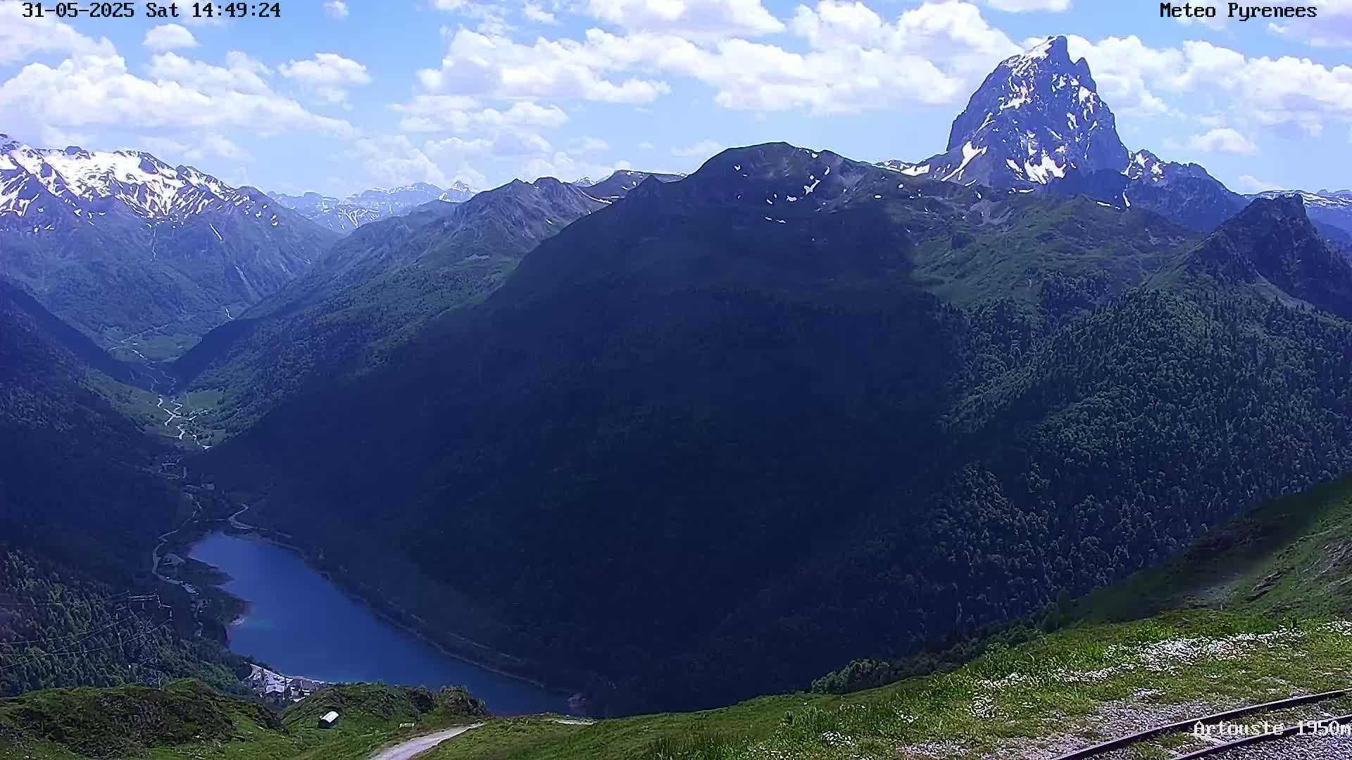 A partly cloudy sky overlooks a mountainous valley containing a lake, with a prominent peak in the background.
