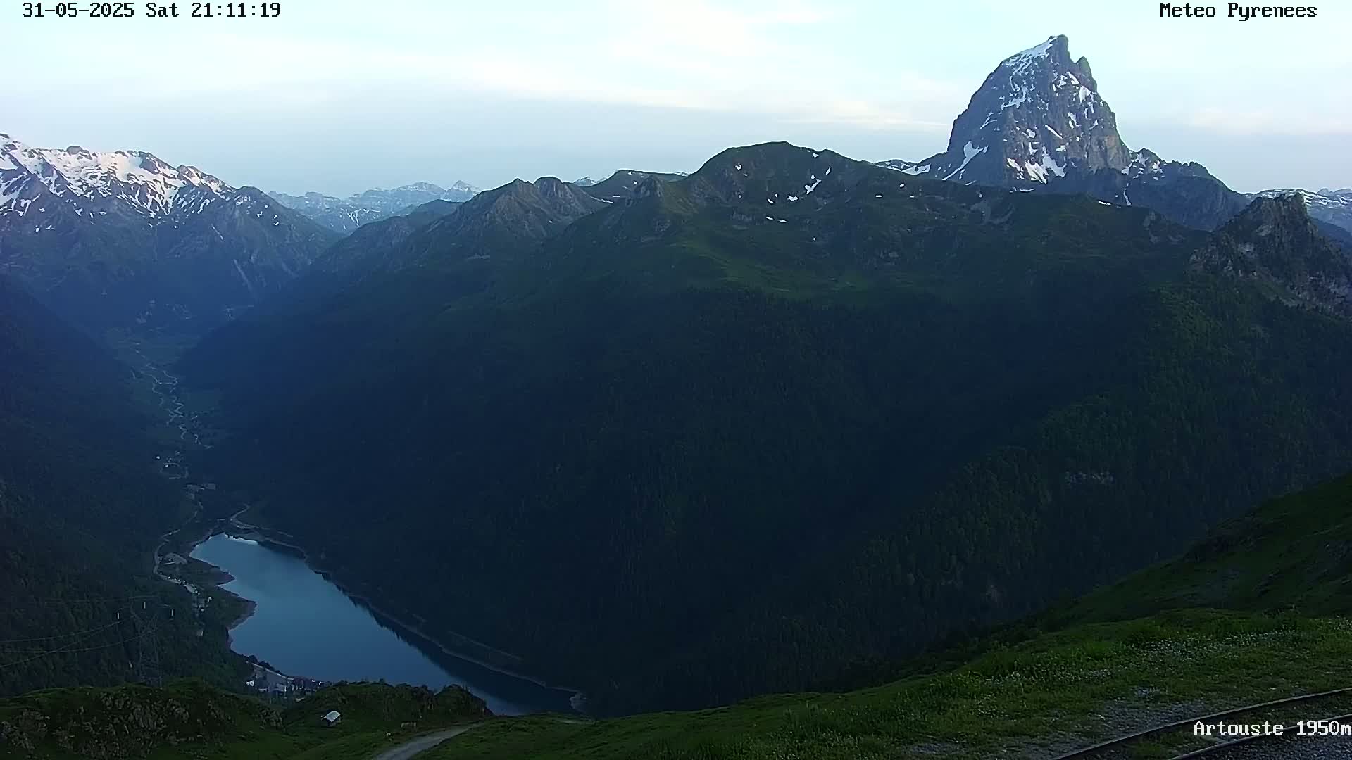 A mountain lake nestled in a valley between dark green mountains, with a prominent, snow-capped peak in the background under a twilight sky.
