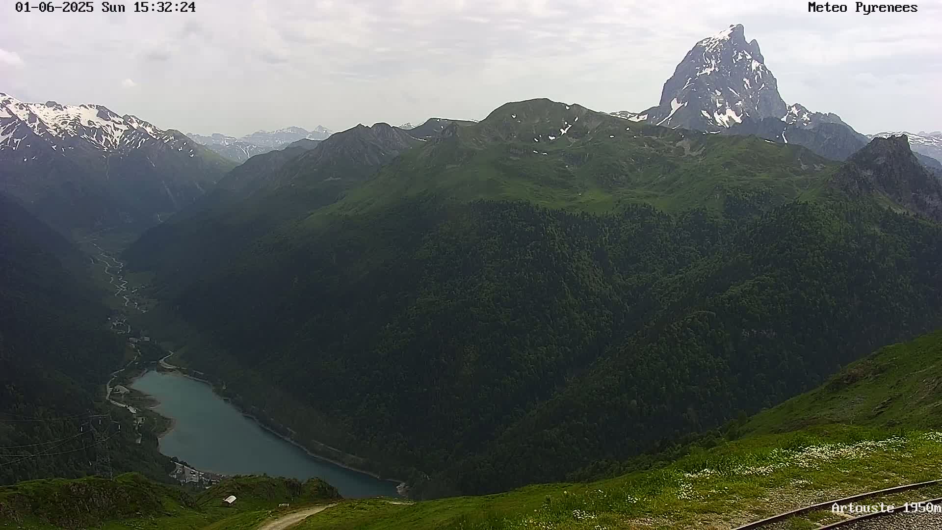 A mountain lake nestled in a valley surrounded by dark green, forested mountains under a cloudy sky.