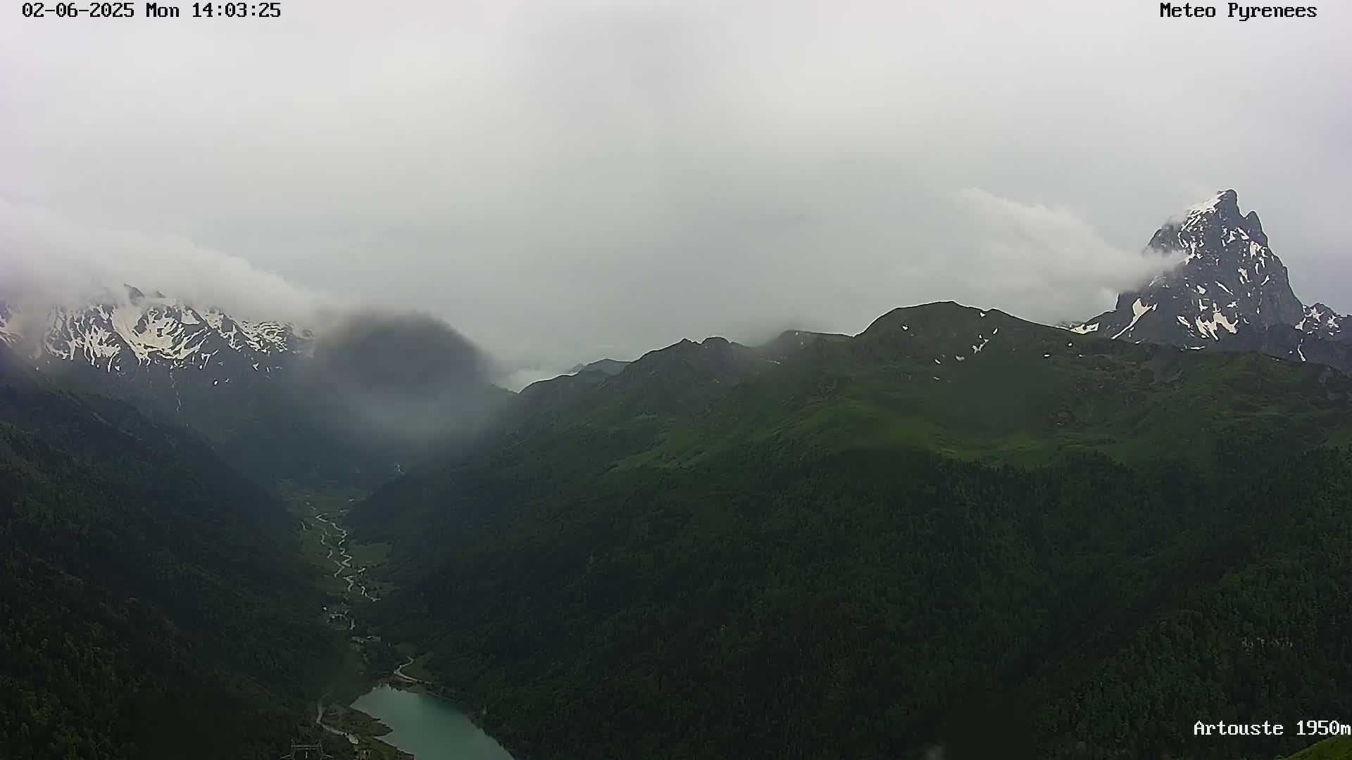 A misty, overcast mountain range with a partially visible lake in a valley below, featuring snow-capped peaks and lush green slopes.