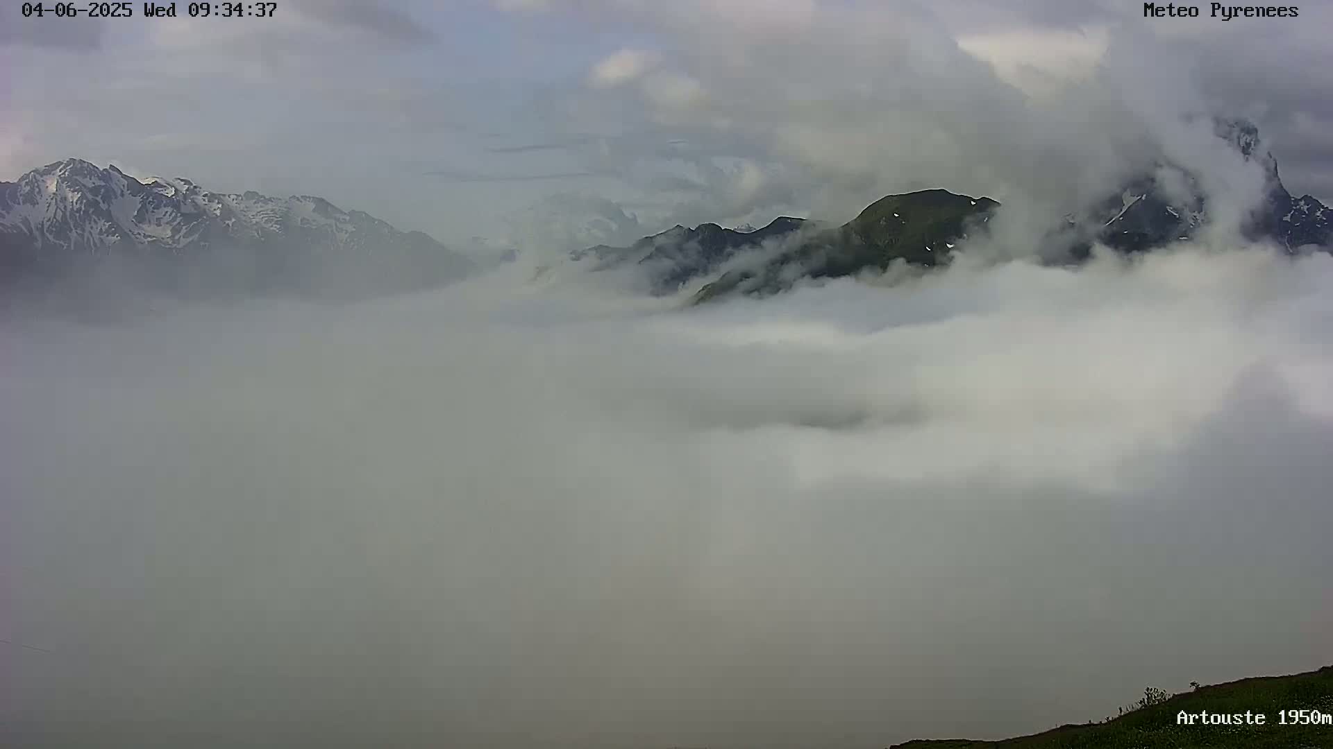 Snow-capped mountains rise above a thick layer of fog under a cloudy sky.