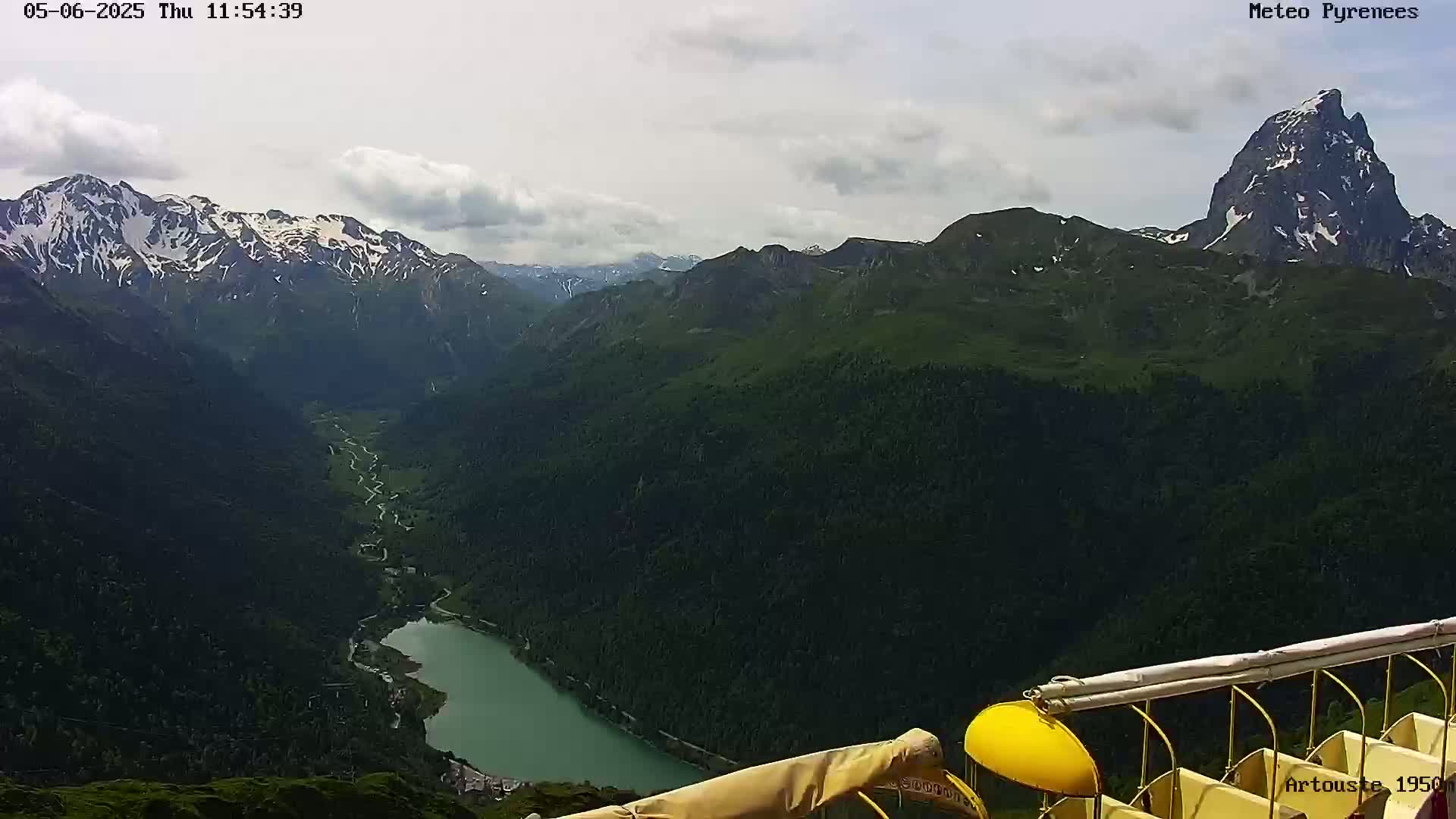 A partly cloudy day reveals a mountainous landscape with a valley containing a lake, viewed from a high vantage point near what appears to be a cable car.