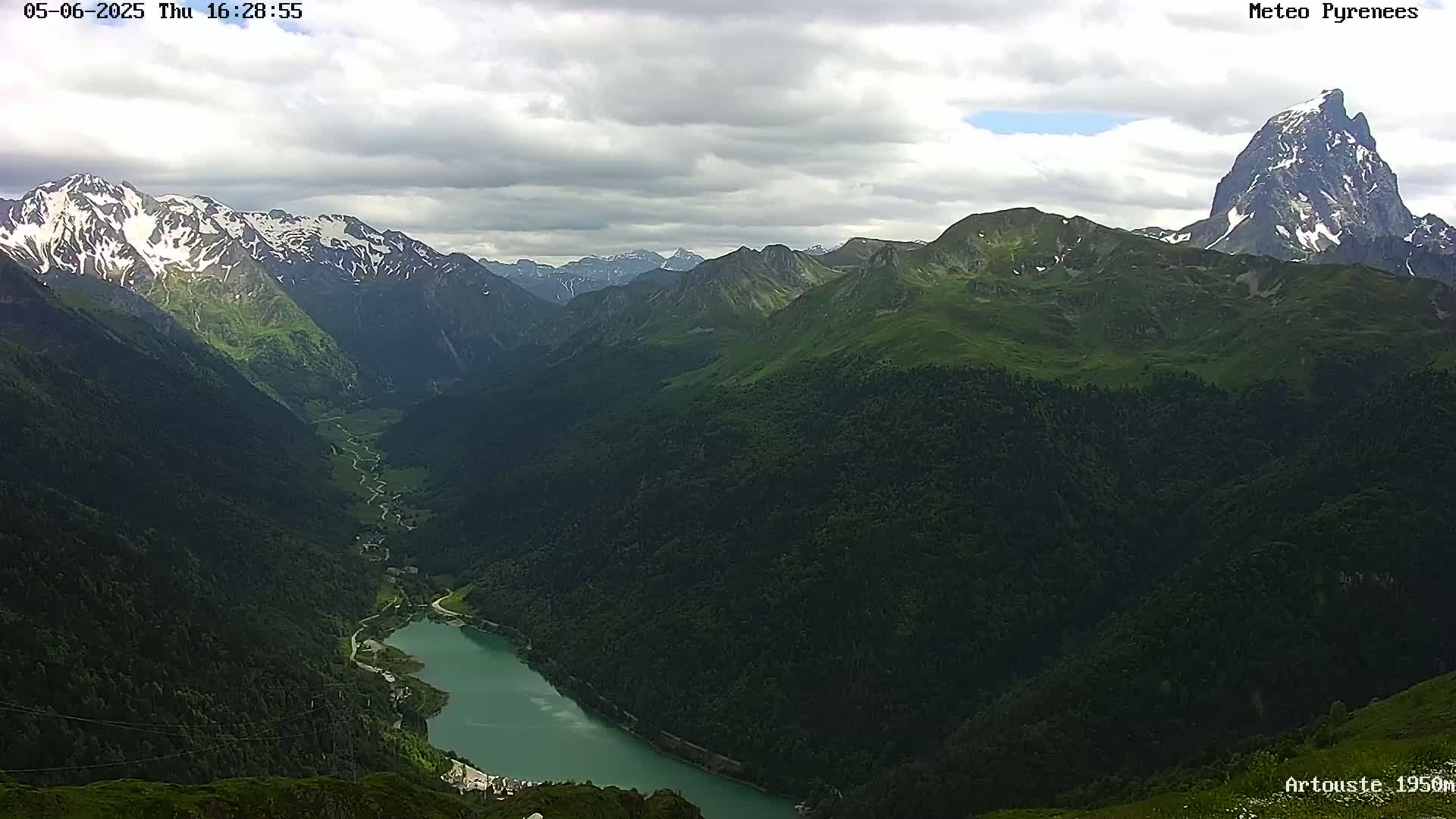 A mountain valley with a teal lake is viewed from above on a partly cloudy day, showing snow-capped mountains in the distance.