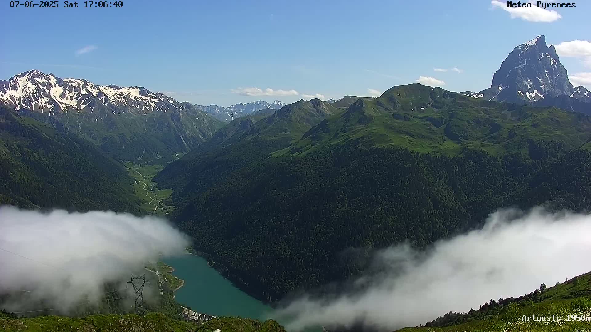 A valley nestled between snow-capped and verdant mountains is partially obscured by low-lying clouds under a clear blue sky.