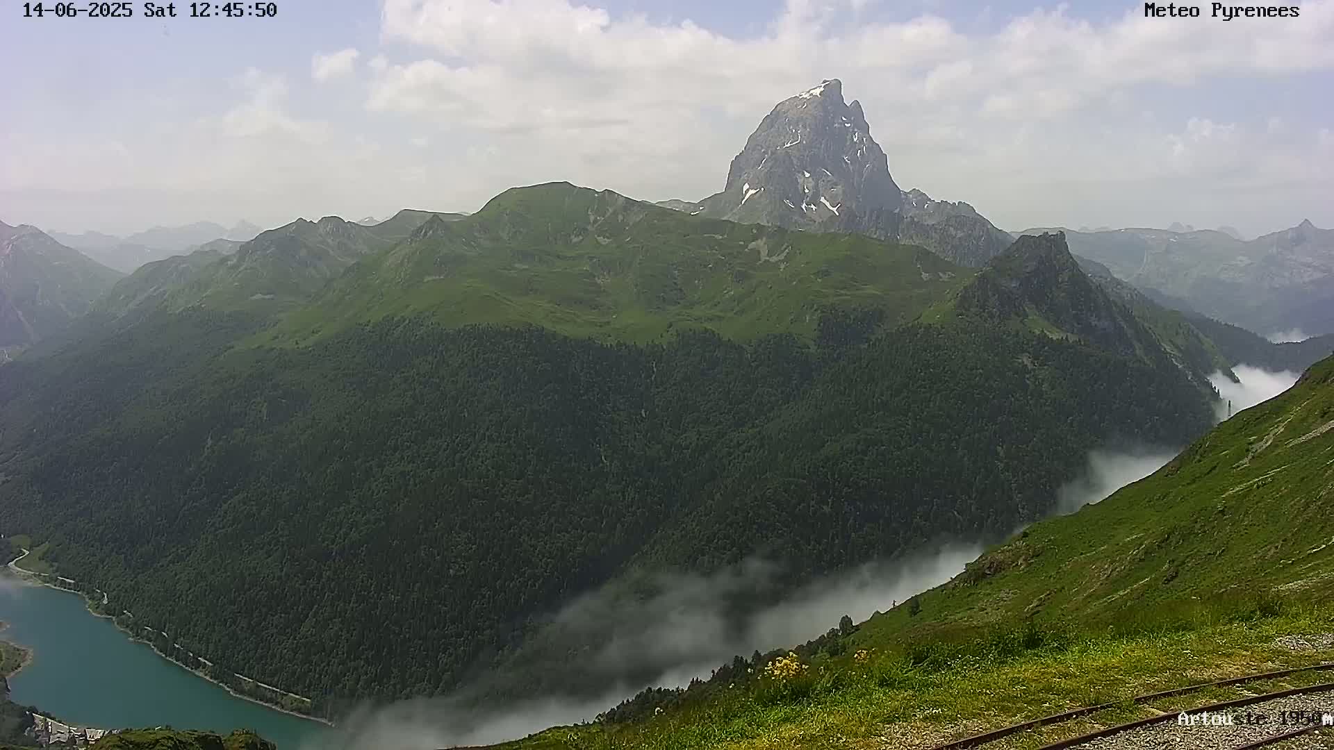 A partly cloudy day reveals a mountainous landscape with a prominent, snow-capped peak, a valley filled with low-lying clouds, and a dark blue lake nestled in the mountains.
