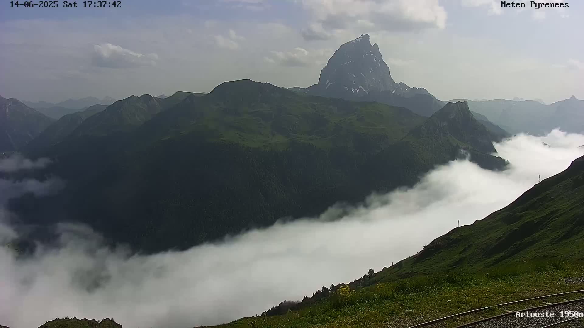 A tall, rocky mountain peak rises above a valley filled with low-lying clouds under a partly cloudy sky.