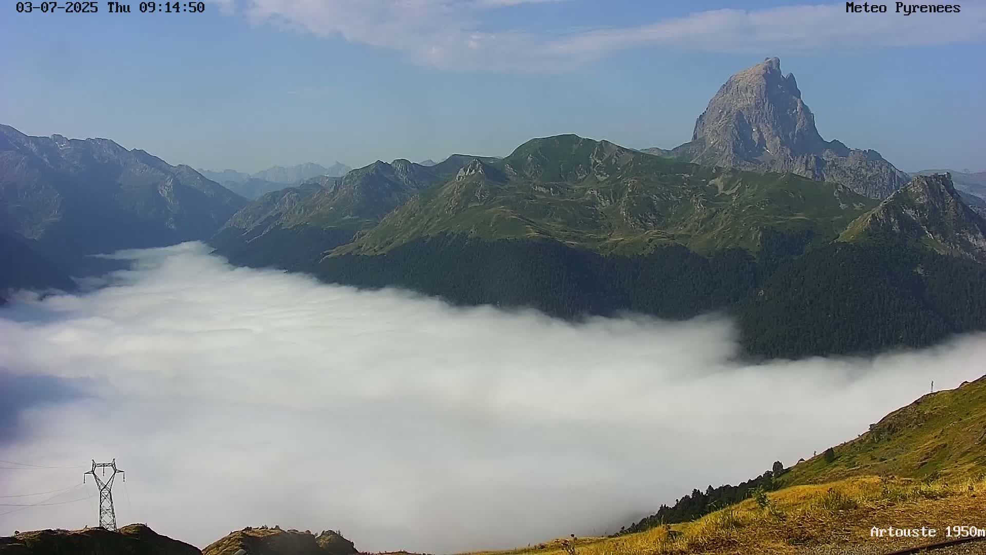 A valley filled with low-lying clouds is nestled between a range of green mountains under a clear sky.