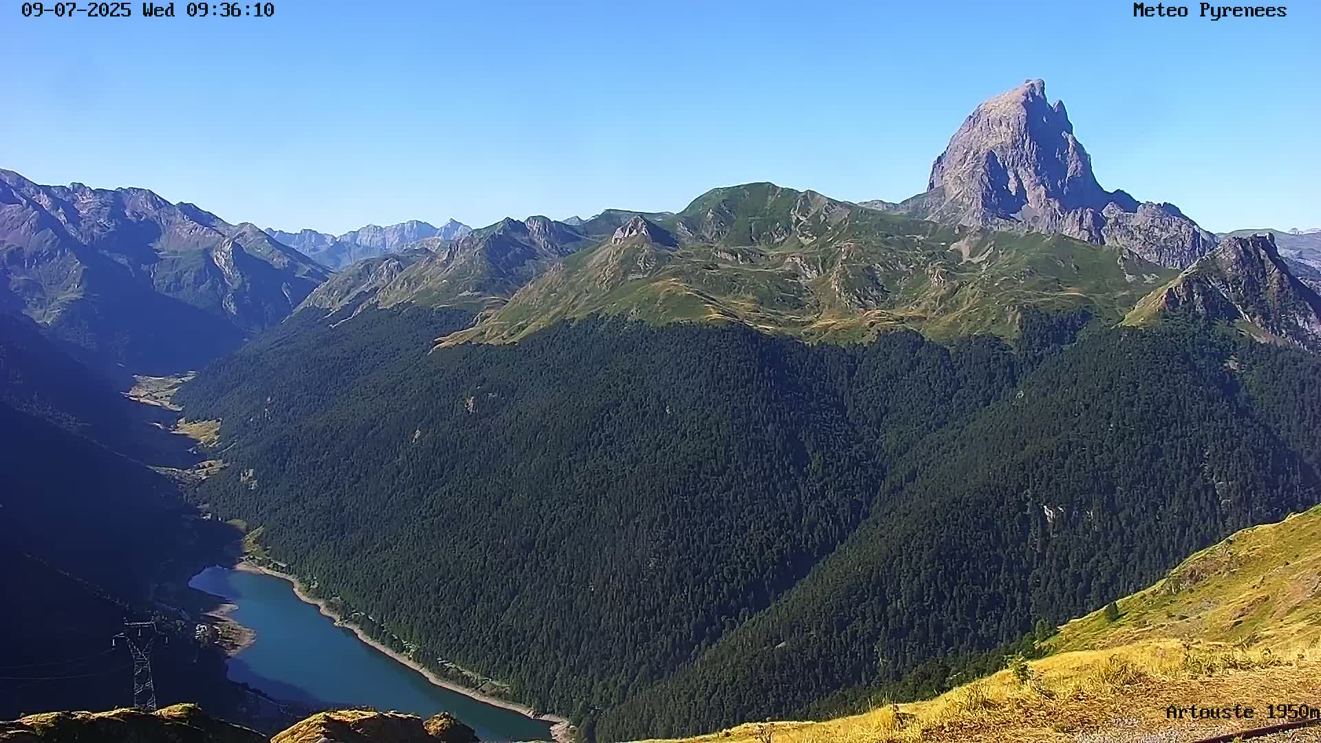 A mountain lake nestled in a valley surrounded by forested mountains under a clear blue sky.