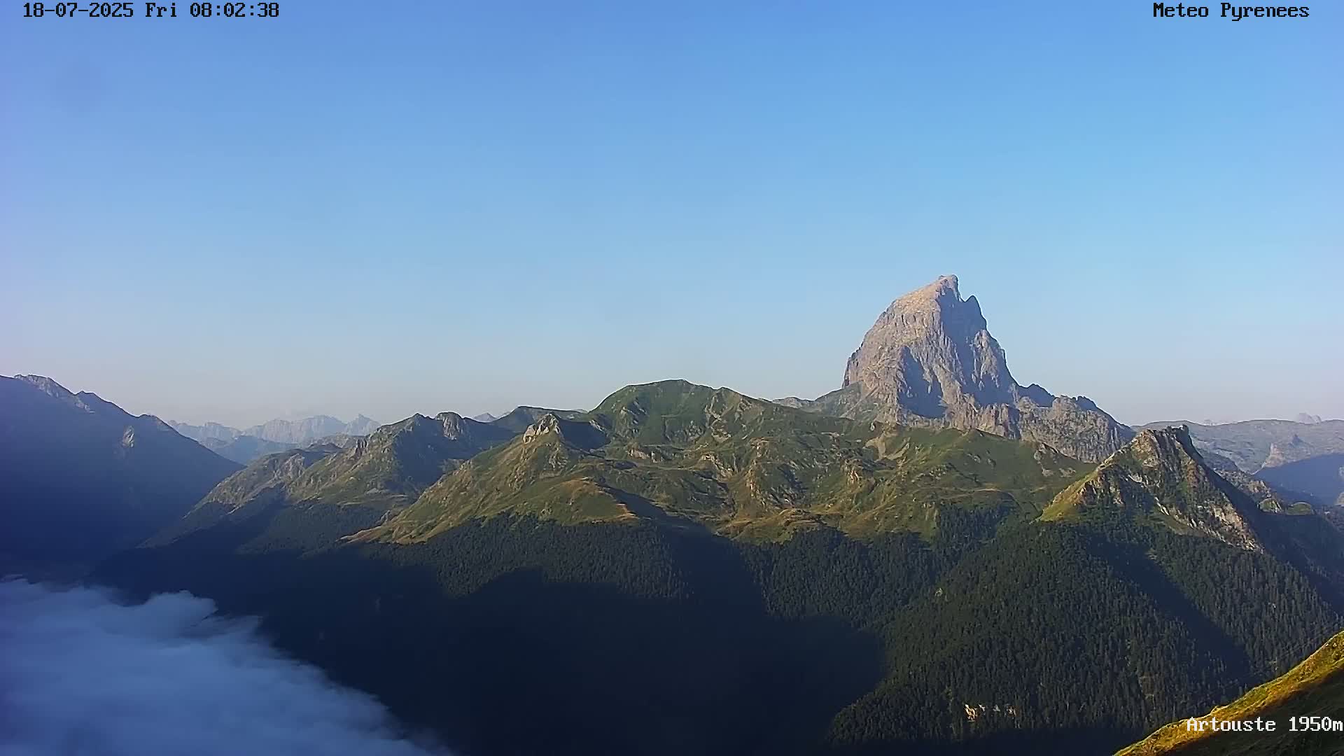 A clear, sunny day reveals a vast mountain range, with a prominent, sharp-peaked mountain in the center, partially obscured by low-lying clouds in the foreground.
