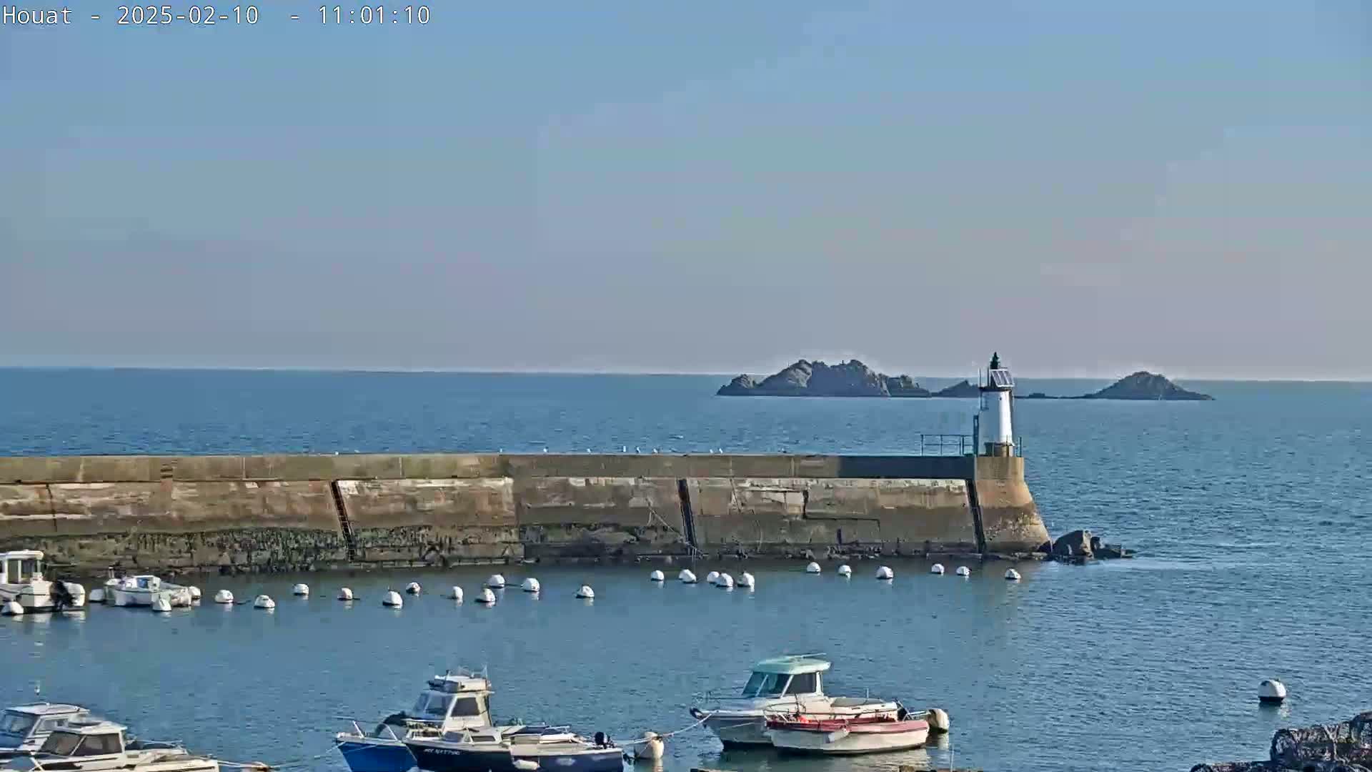 A harbor with several small boats is partially enclosed by a long, low stone wall, beyond which a small lighthouse stands on a rocky outcrop in calm, clear blue water.