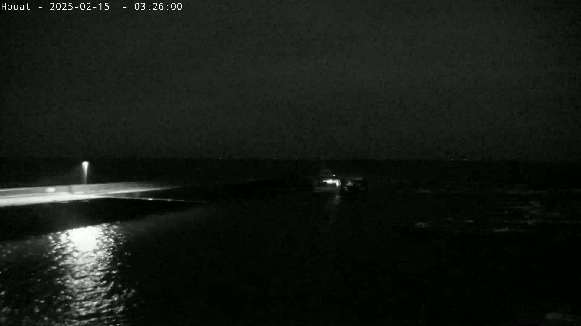 A nighttime, low-light view shows a dark body of water with a lit roadway and a small vessel in the distance.