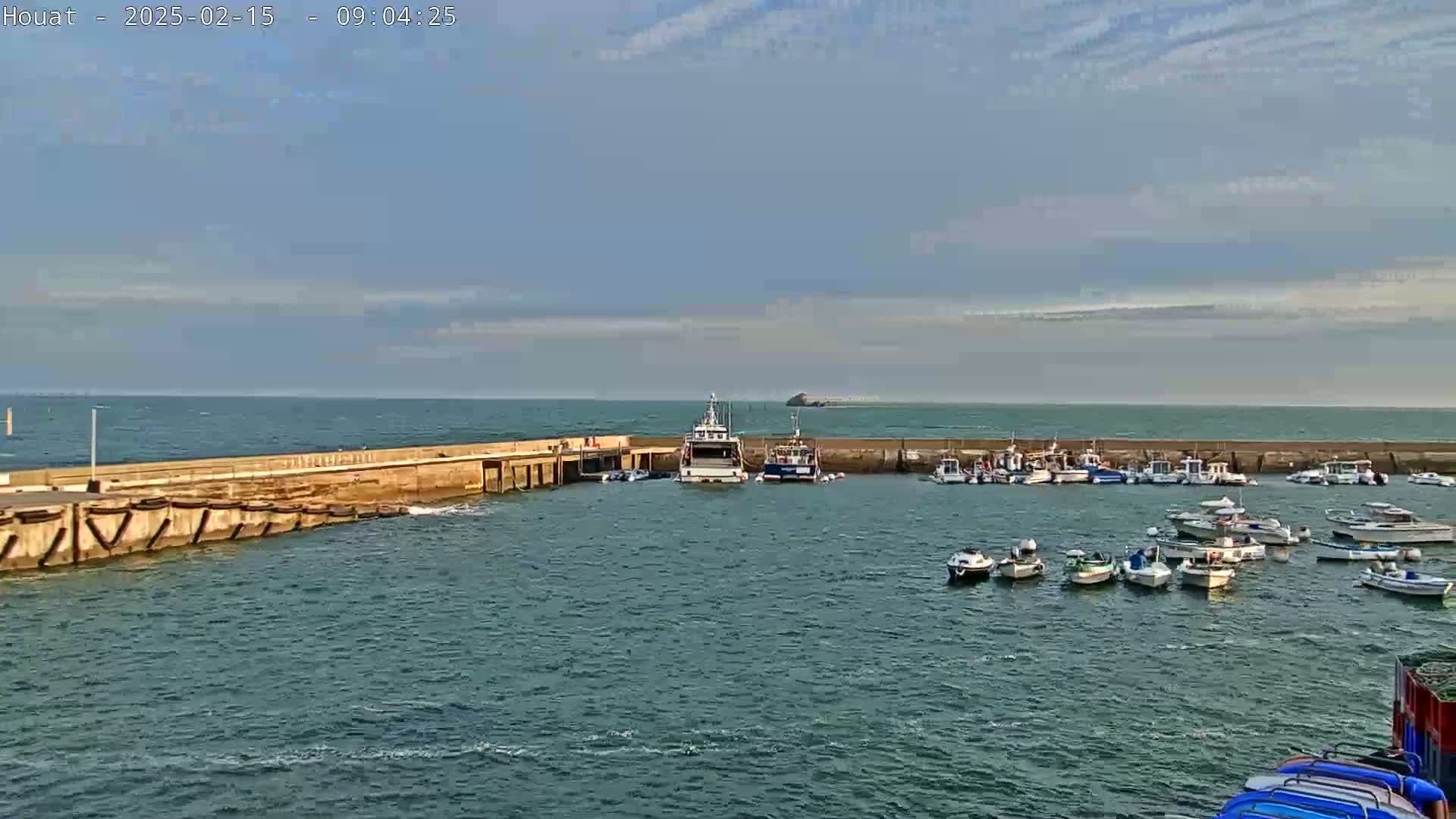 A harbor filled with numerous small boats is partially sheltered by a concrete seawall under a mostly clear sky.
