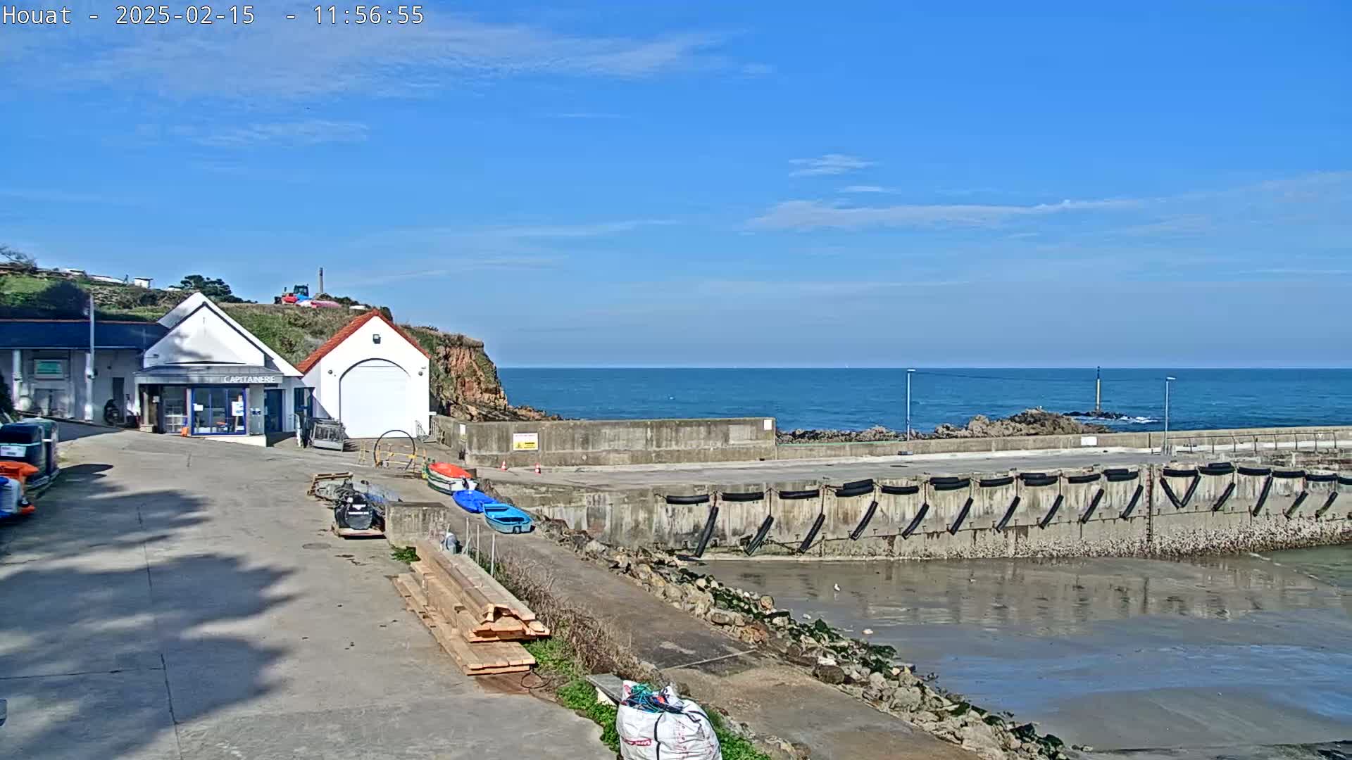 A small harbor with a building, several small boats, and a mostly calm ocean under a clear blue sky.