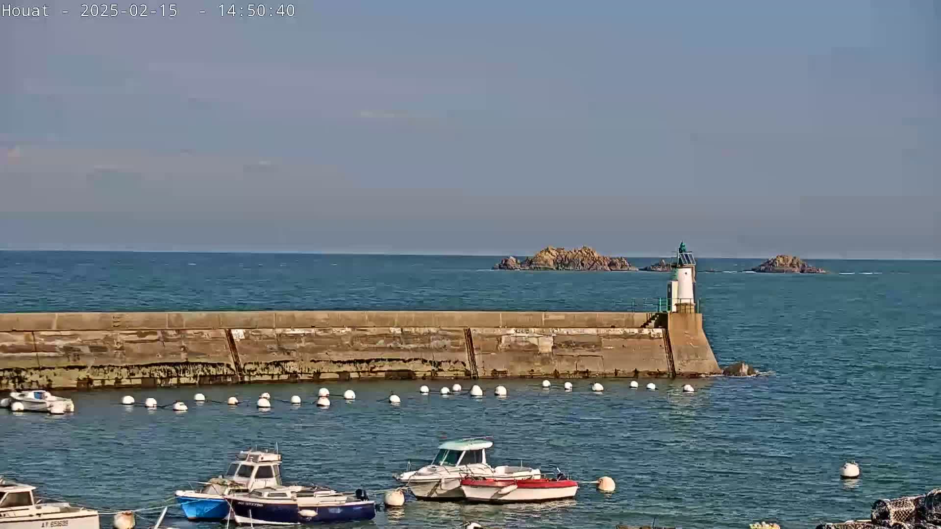 Several small boats are moored in a harbor protected by a stone seawall and a lighthouse, with calm seas and clear skies in the background.