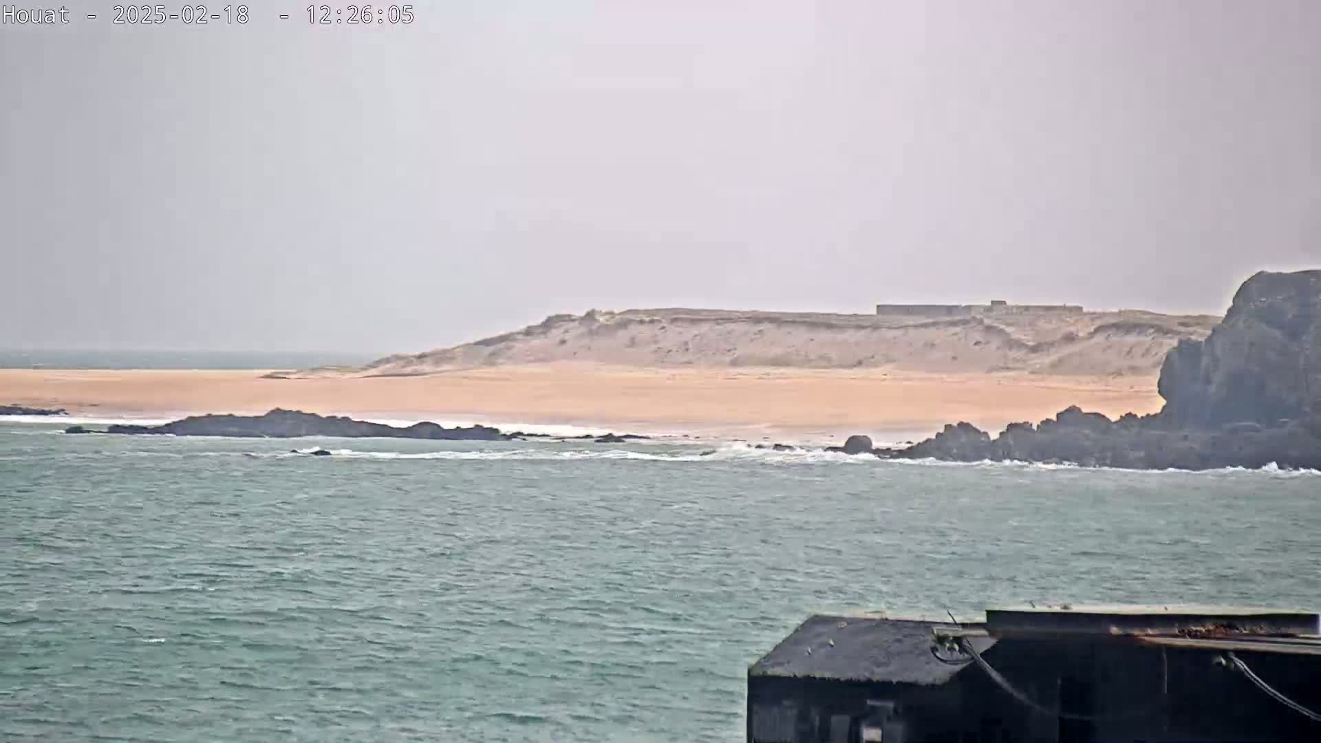 A sandy beach backed by dunes and dark rocks is visible across a calm, light-green body of water, under an overcast sky.