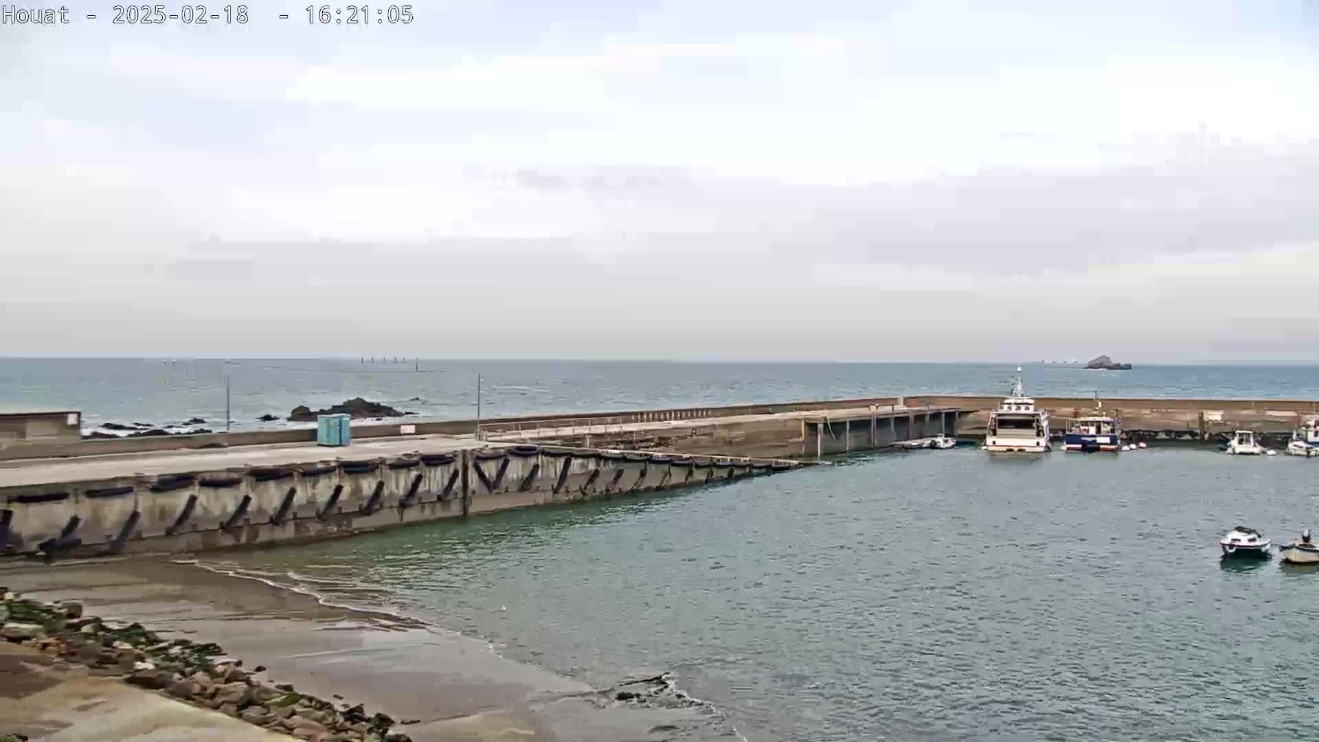 A small harbor with several boats is seen on an overcast day, next to a concrete seawall that extends into a calm body of water.