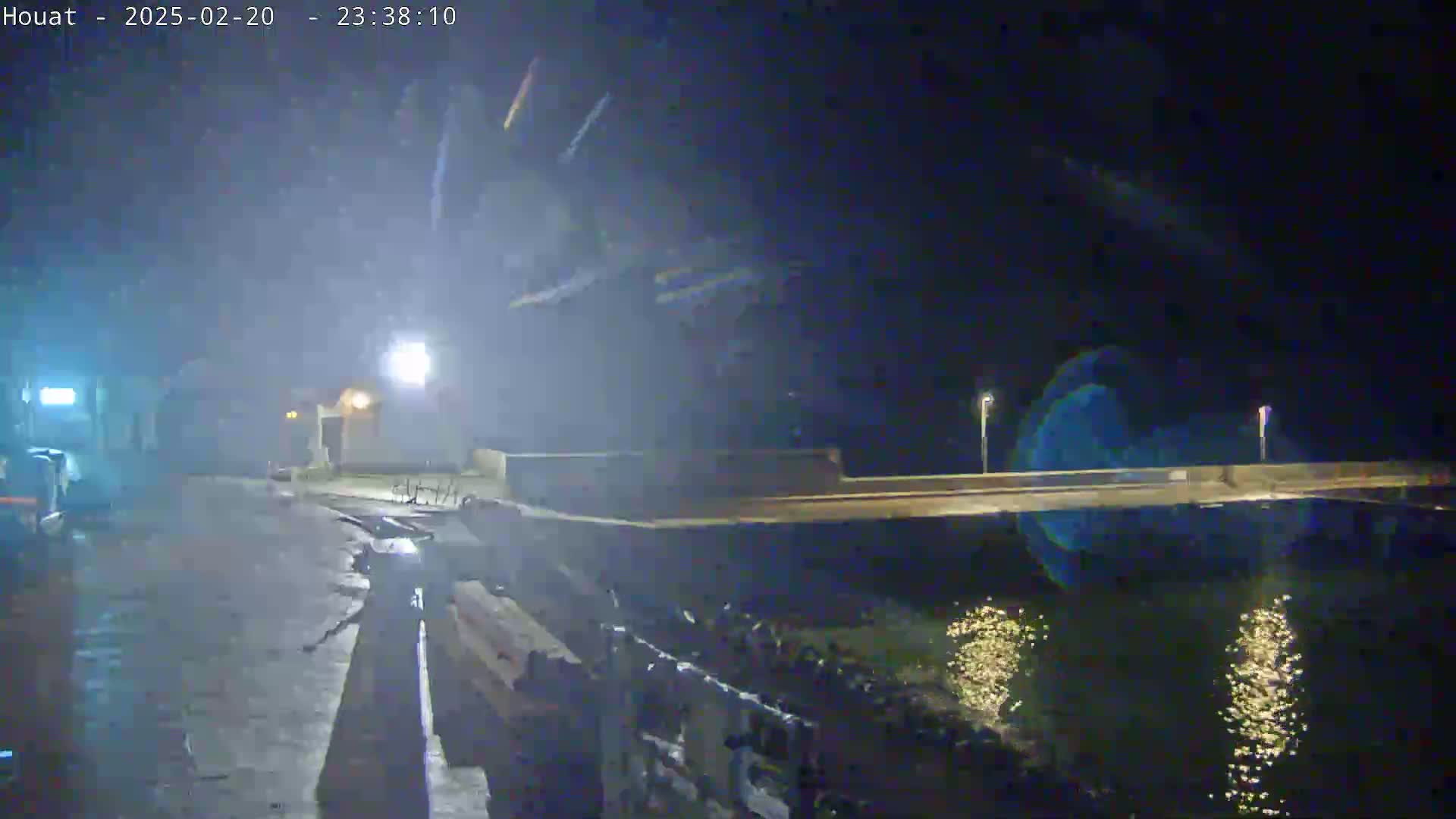 A nighttime, possibly rainy, scene shows a low-lit road leading to a bridge over water, with lights reflecting on the water's surface.