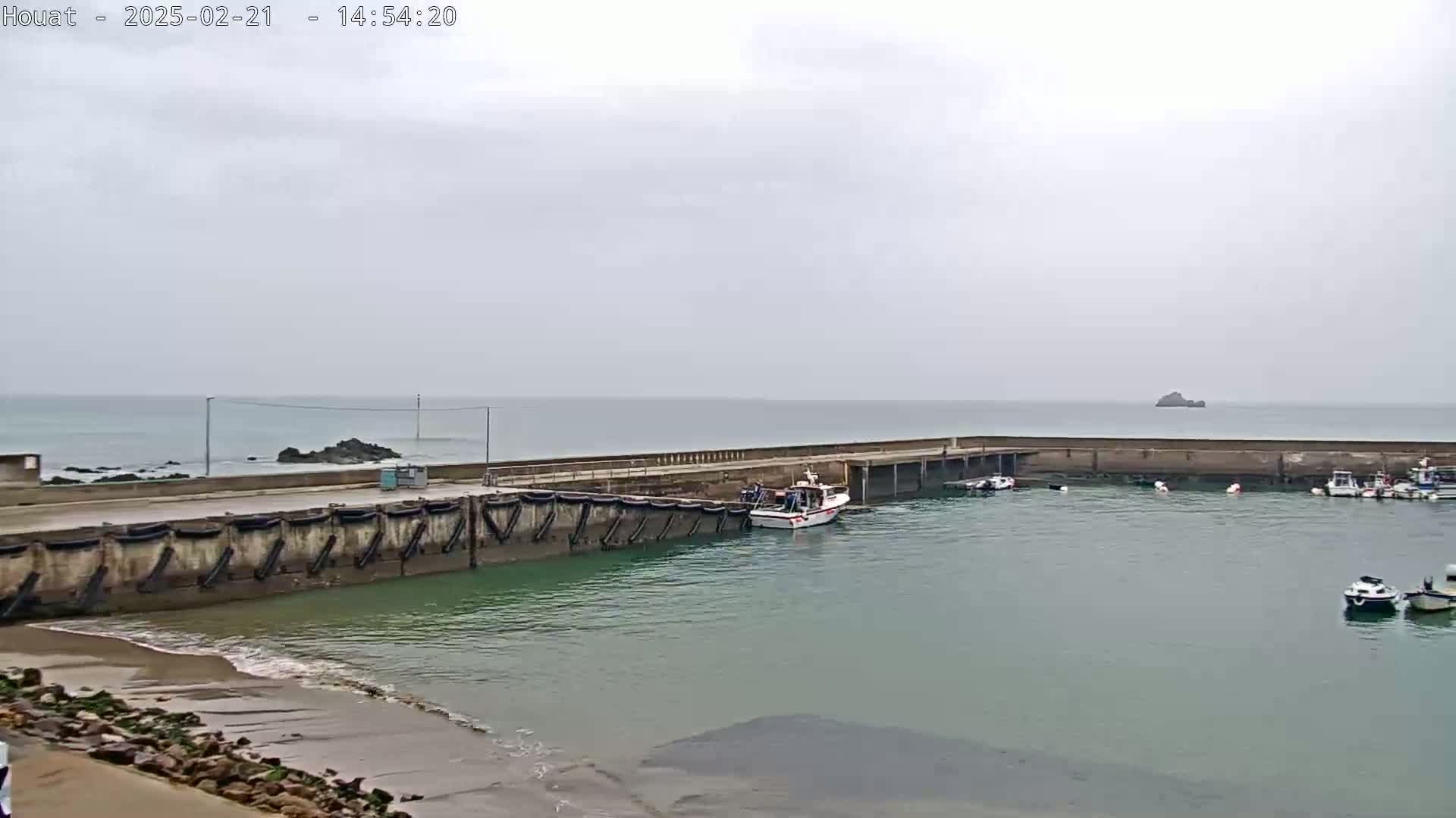 A small harbor with several boats is seen on an overcast day, with calm water and a rocky outcrop visible in the distance.